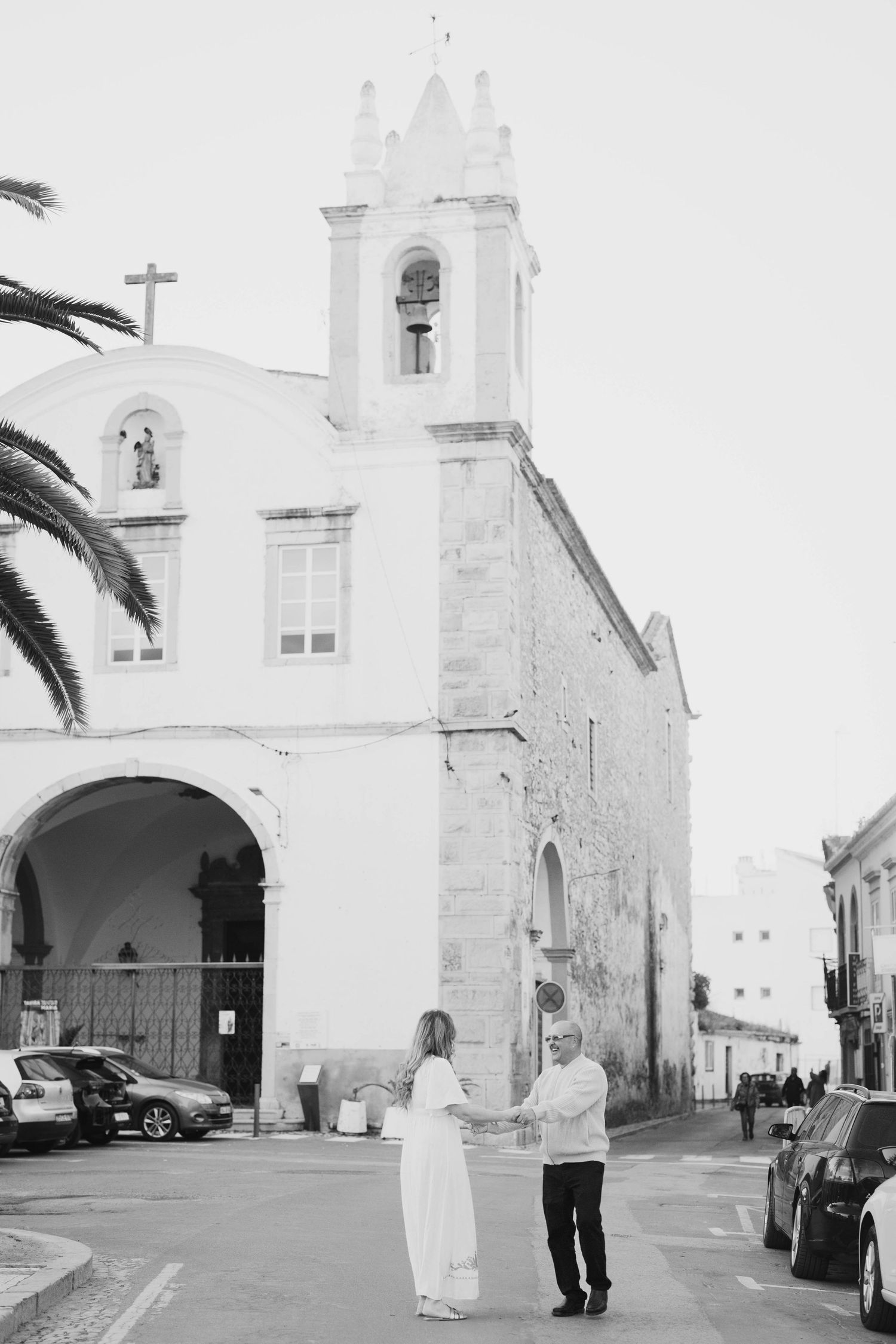Black and white photo of a couple holding hands in front of a white church with a bell tower and palm tree.