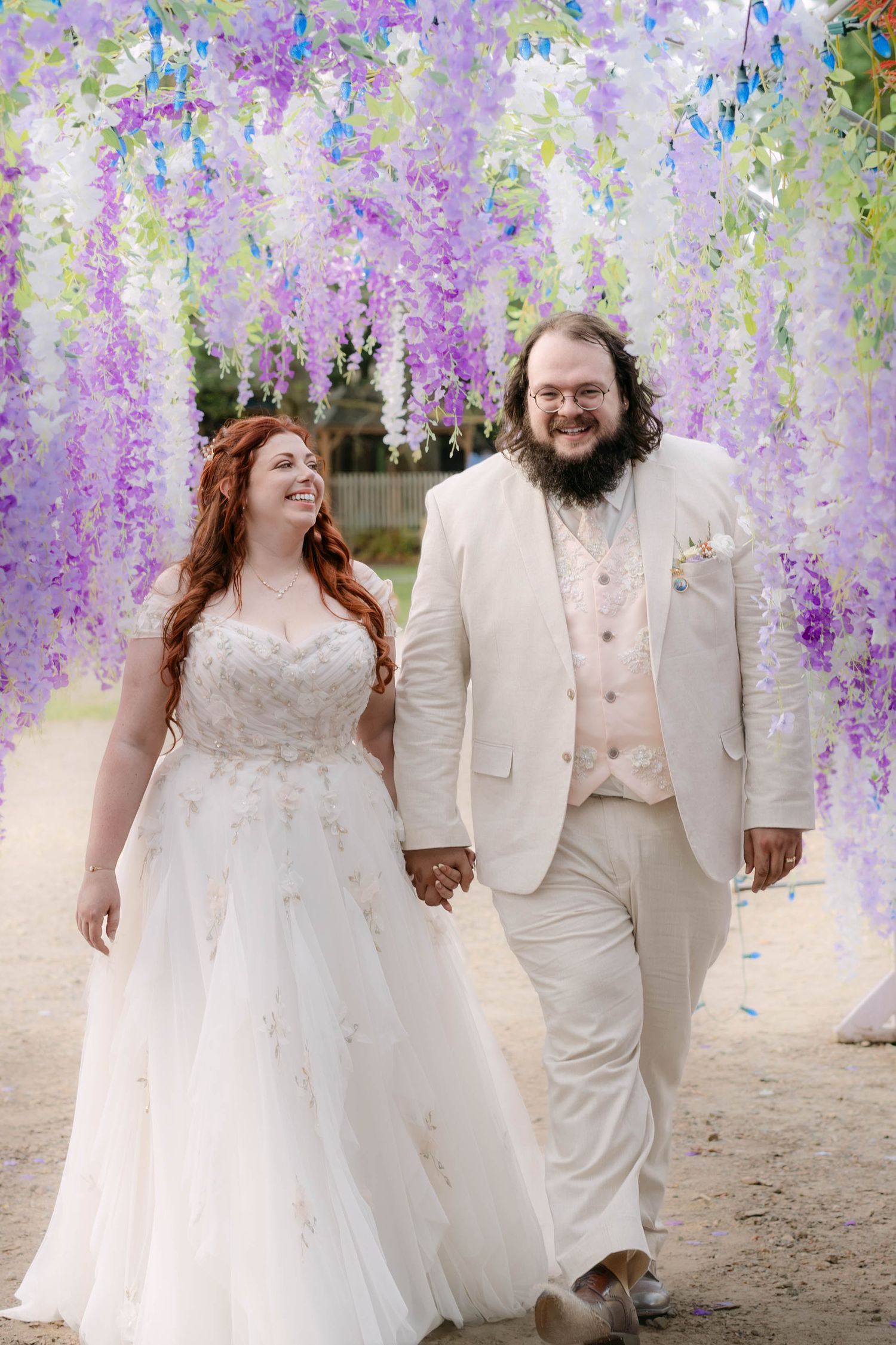 A joyful bride and groom walk hand in hand beneath cascading purple wisteria floral decorations on their wedding day.