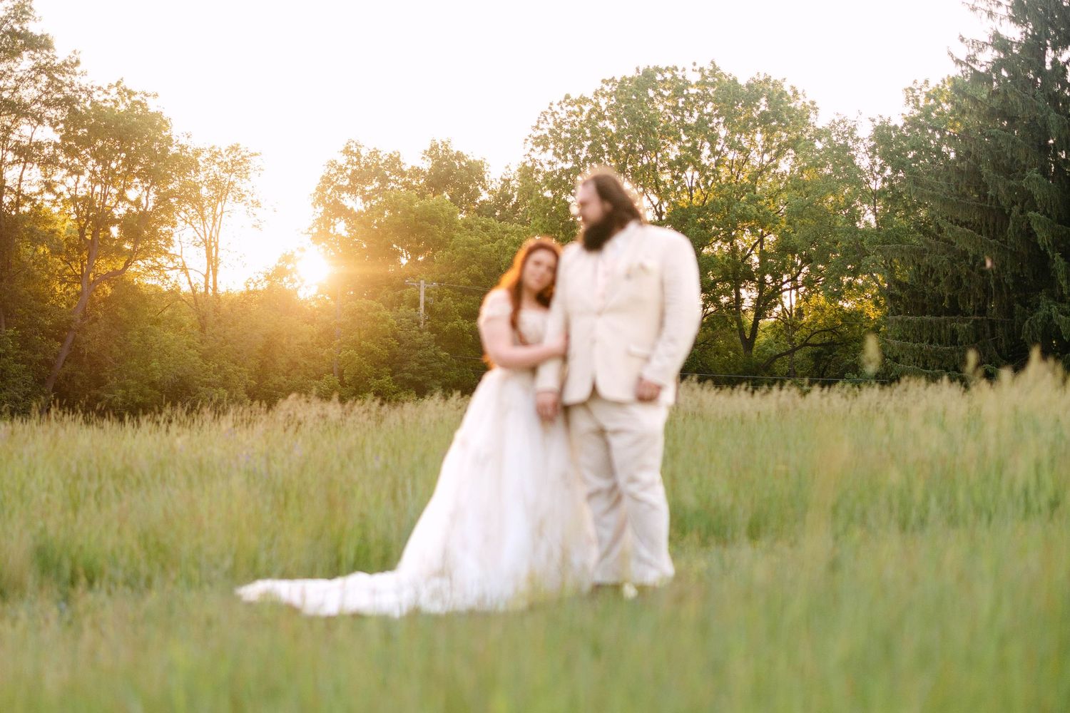Bride and groom stand in golden sunlit meadow, bride in flowing white gown, groom in tan suit, surrounded by lush green trees.