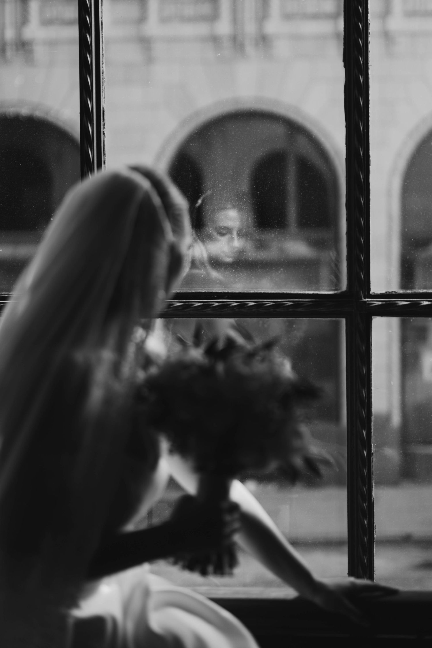 Black and white photo of a bride holding a bouquet, her reflection visible in a vintage window pane.