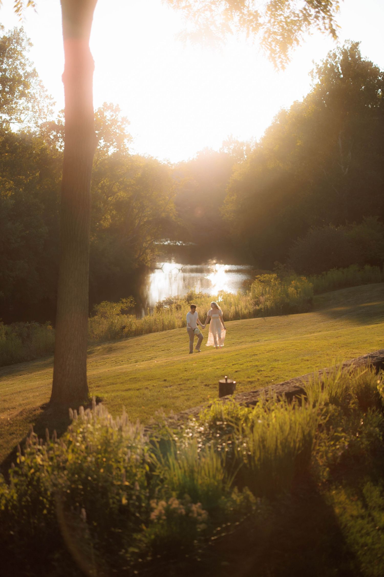 Couple holding hands walking toward a sunlit pond surrounded by lush green trees during golden hour sunset.