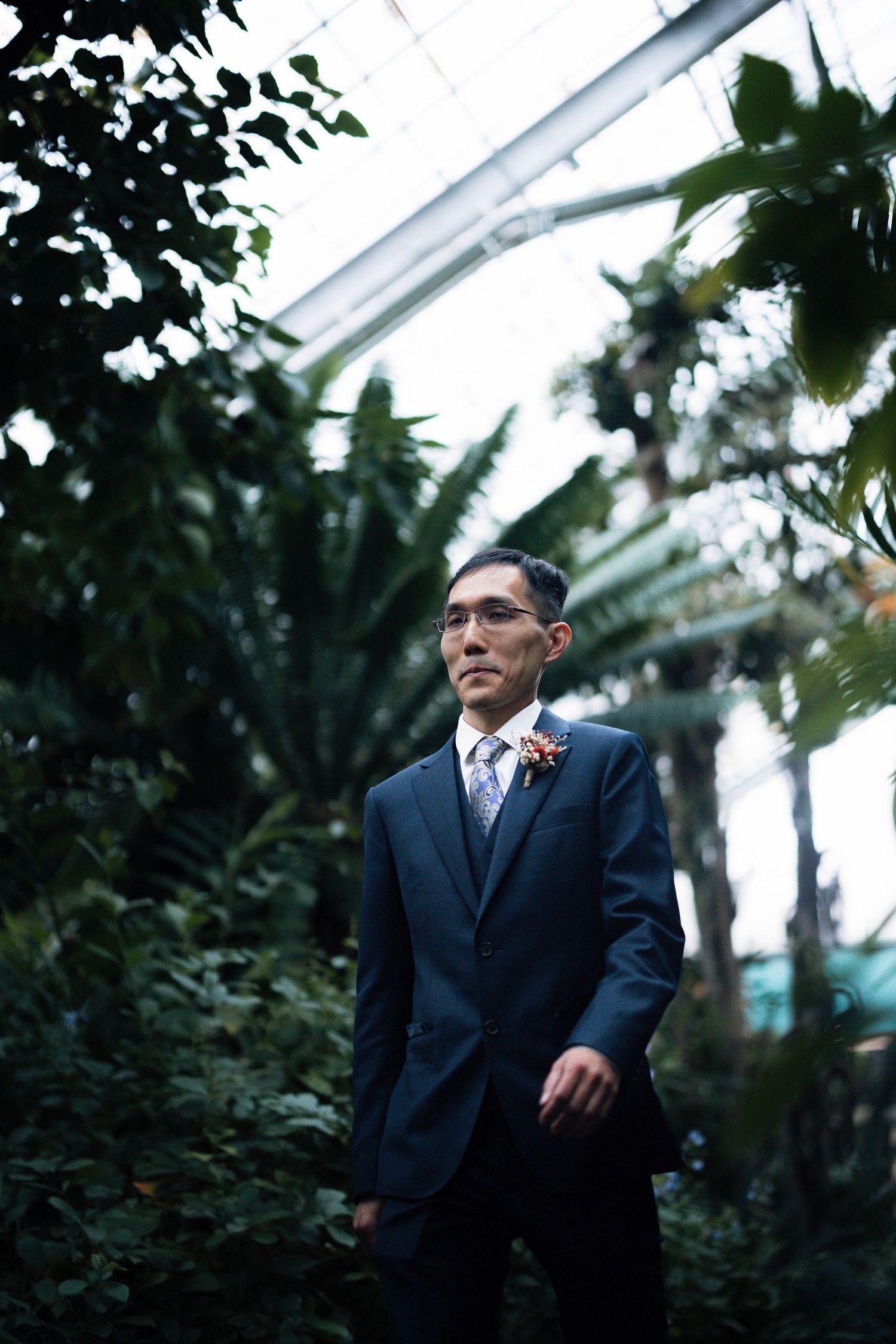 Groom in navy suit with boutonniere standing among tropical plants in greenhouse.
