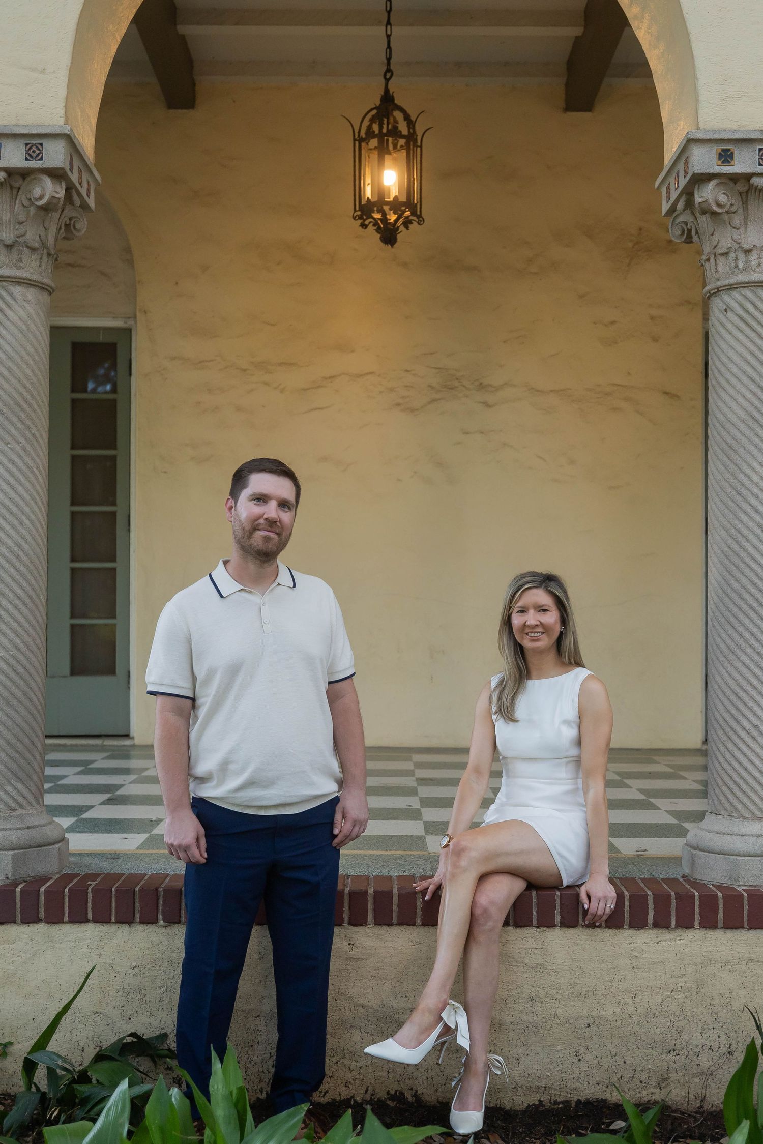 Couple posing under ornate archway with lantern, man in polo shirt and woman in white mini dress.