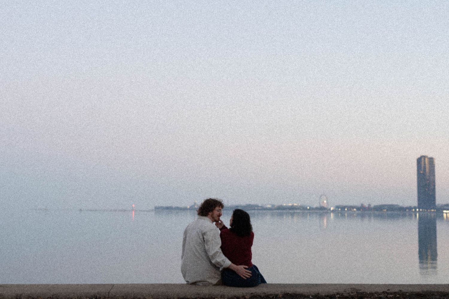 Romantic couple sitting together on a waterfront ledge at dusk, gazing at a calm lake with a city skyline in the distance.
