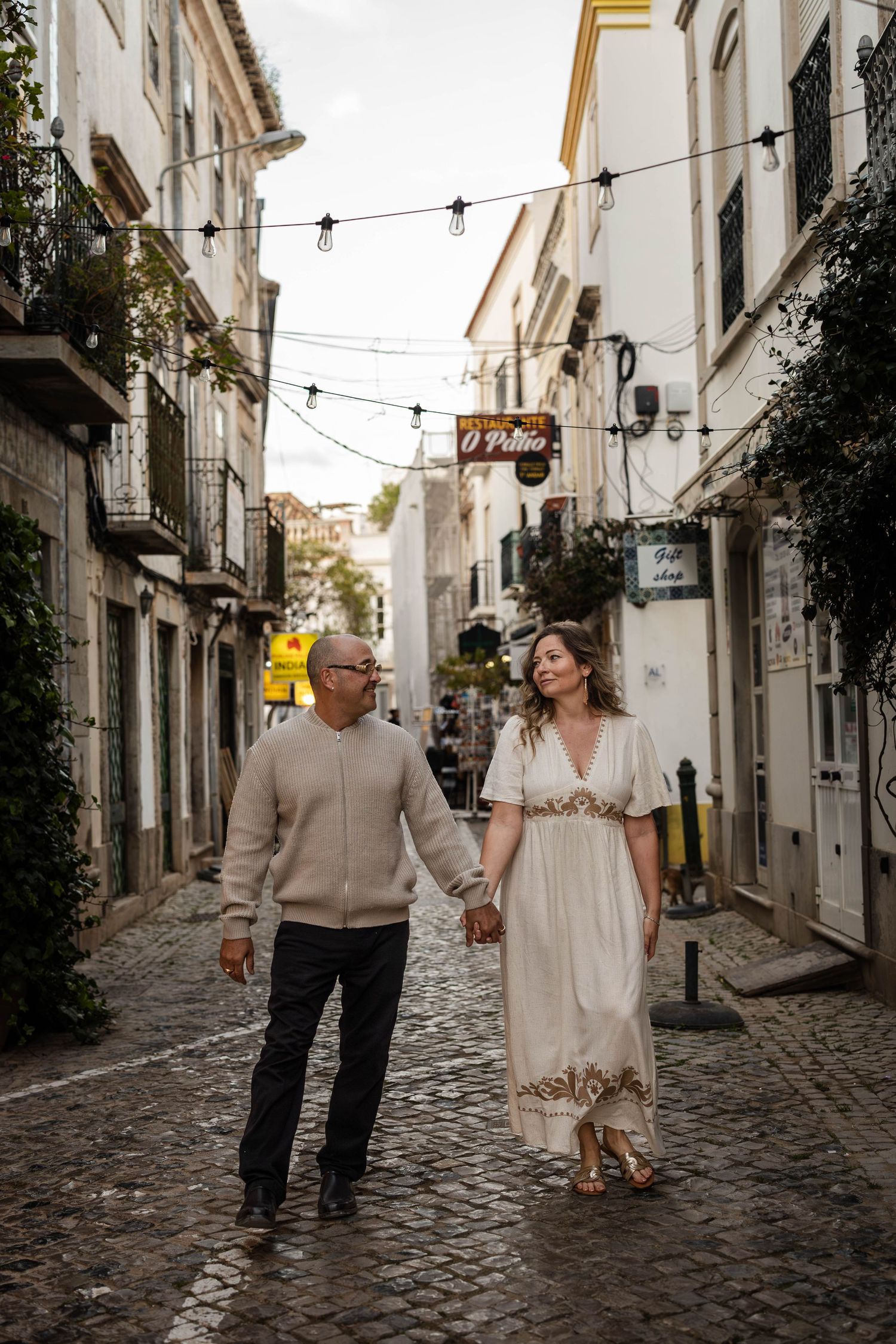 Couple holding hands walking on cobblestone street in historic European town with string lights overhead.