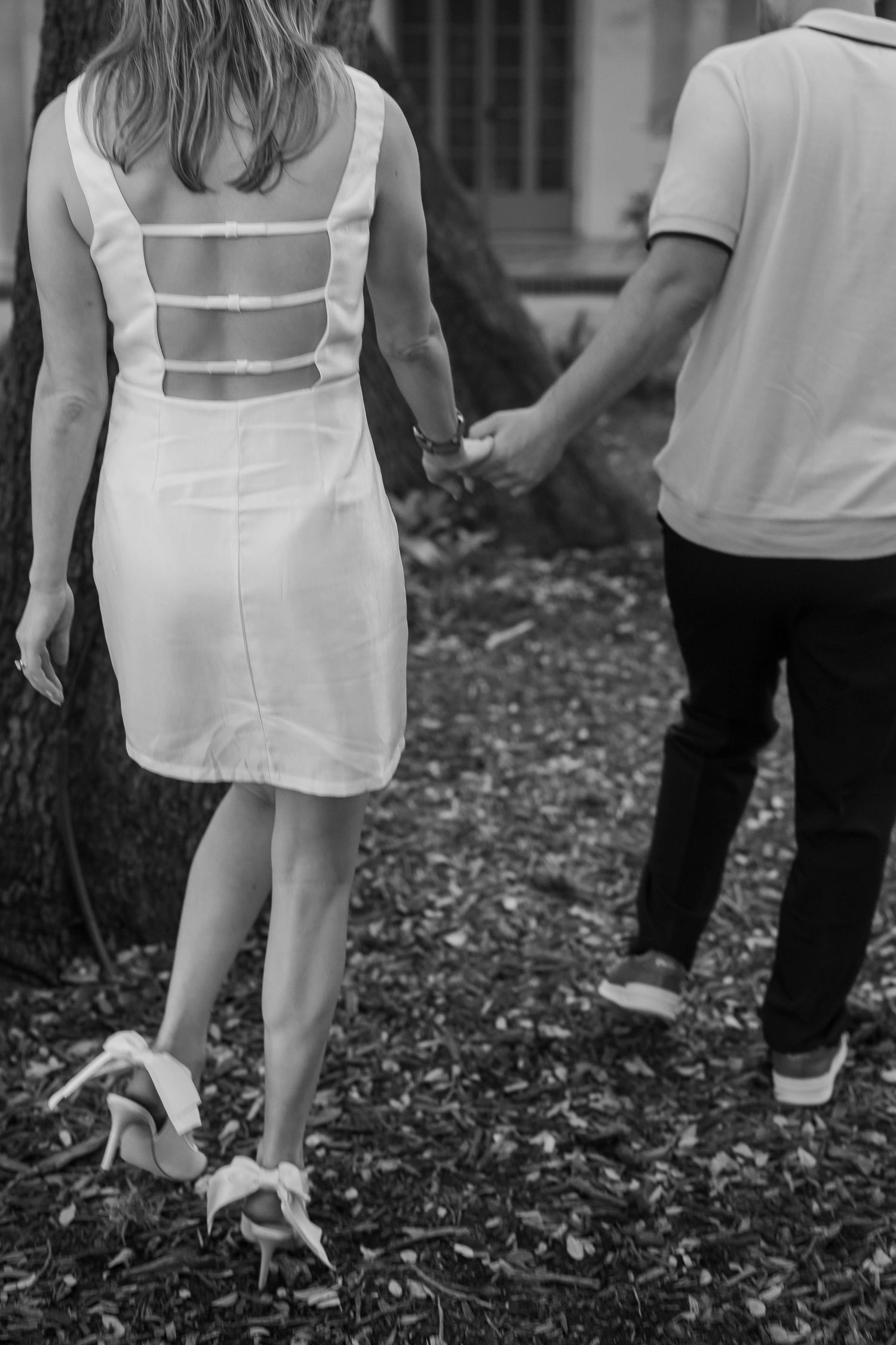 Black and white photo of couple holding hands, woman in white open-back dress and heels walking outdoors.