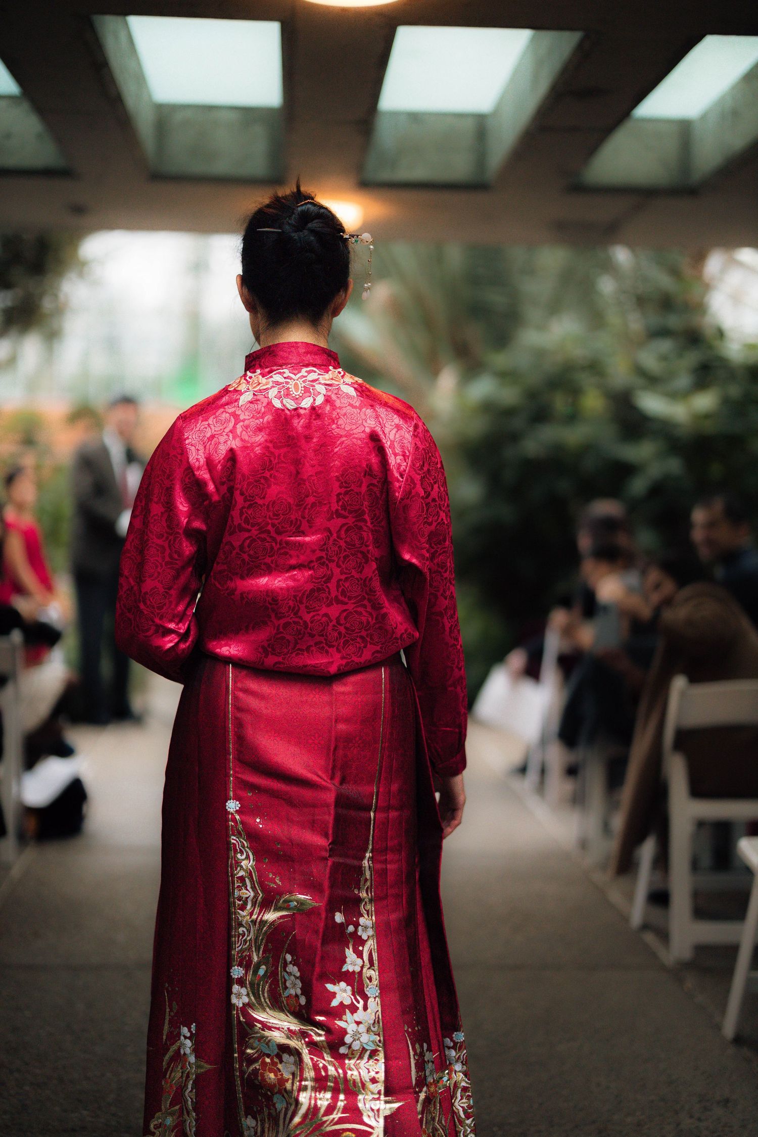 Bride in traditional red Chinese qipao wedding dress walks down aisle in tropical greenhouse venue.