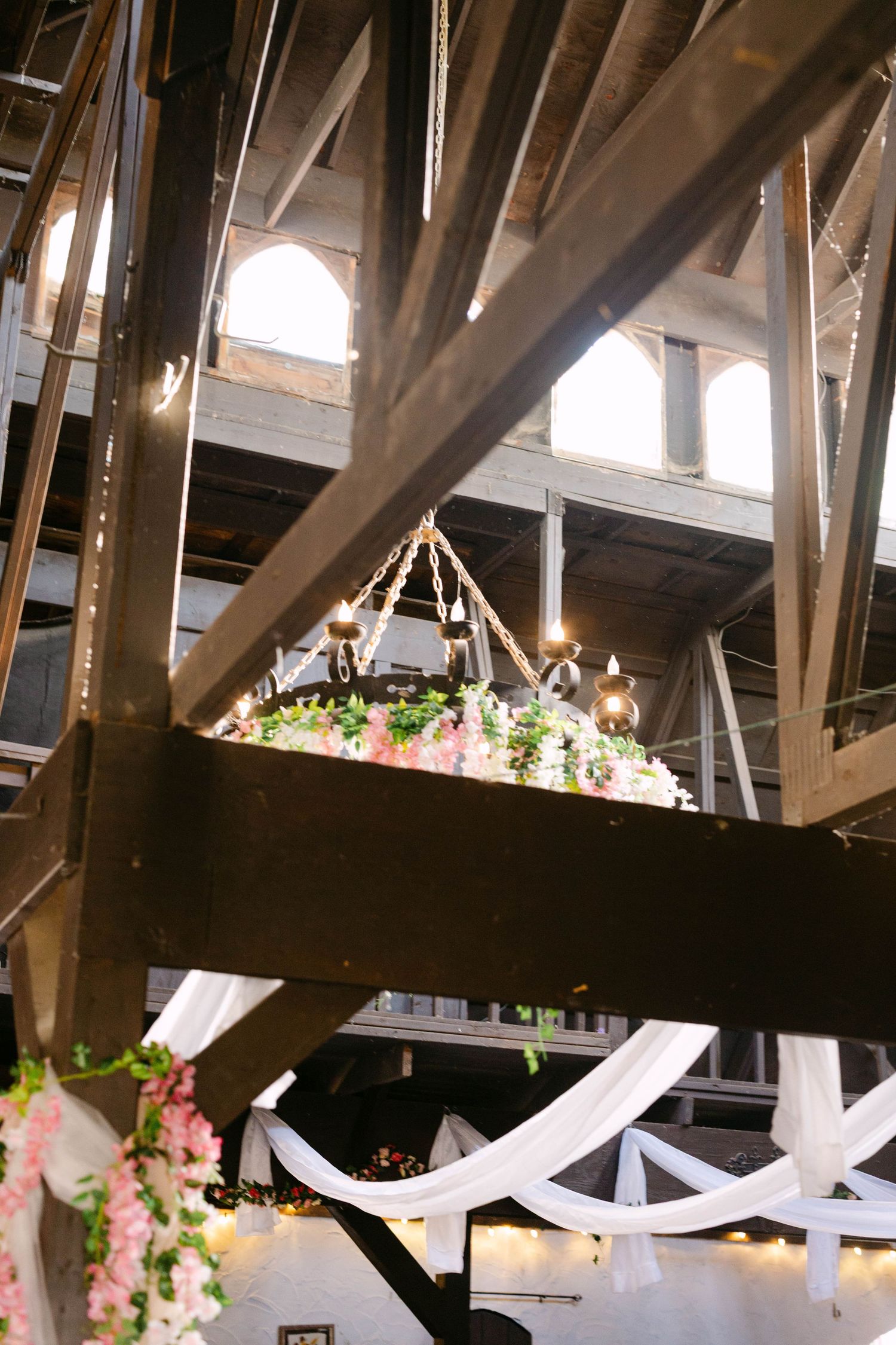 Elegant wedding venue interior with floral chandelier, string lights, white draping, and rustic wooden beam ceiling.