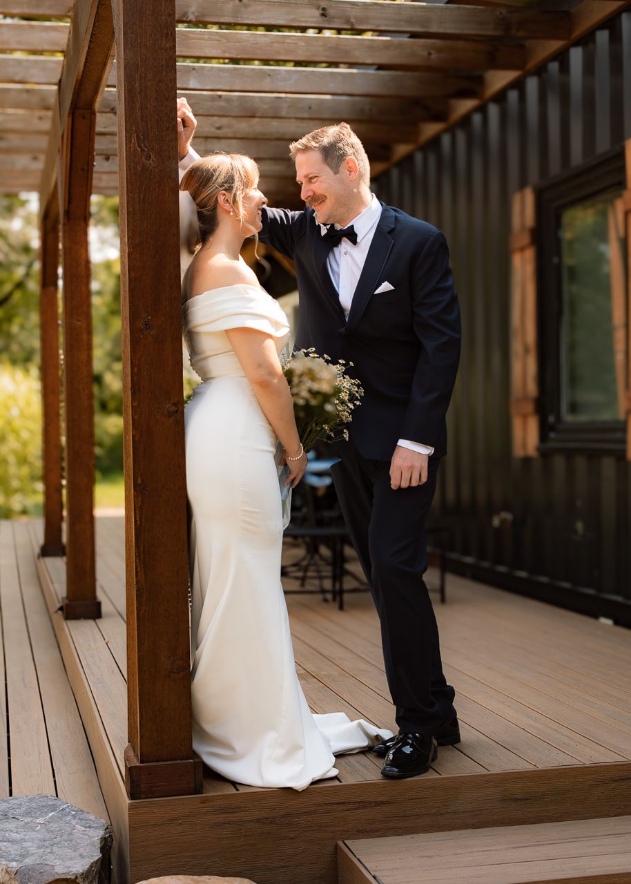 Bride in off-shoulder white gown and groom in black tuxedo share a romantic moment on a wooden deck.