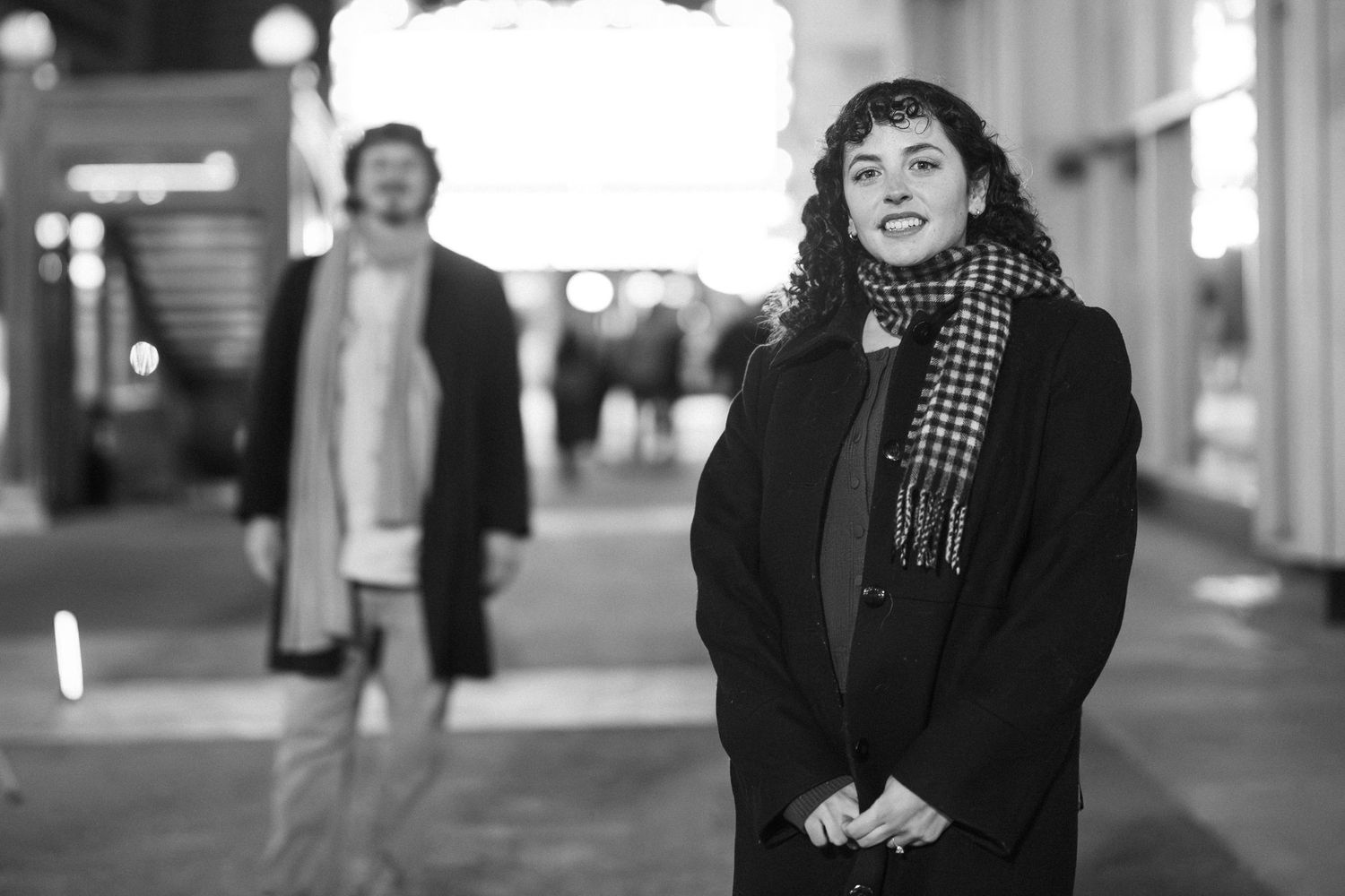 Black and white photo of a woman in a houndstooth scarf smiling on a city street at night.