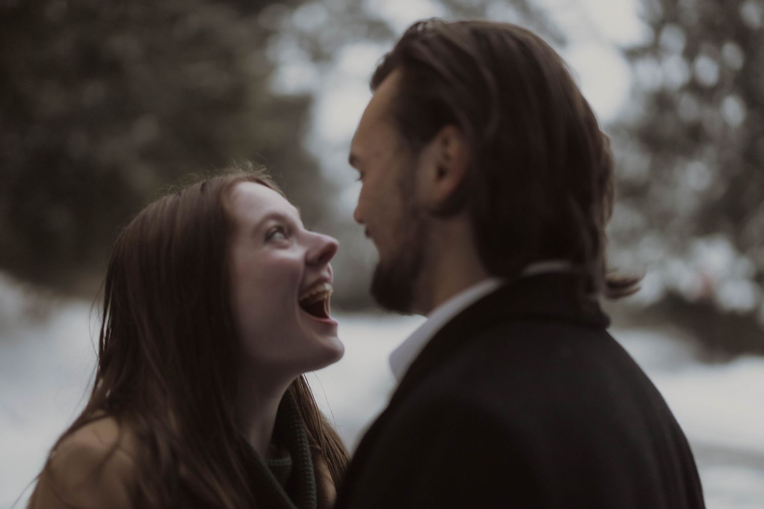 A joyful woman laughs while gazing at a bearded man outdoors in a wintry, romantic setting.