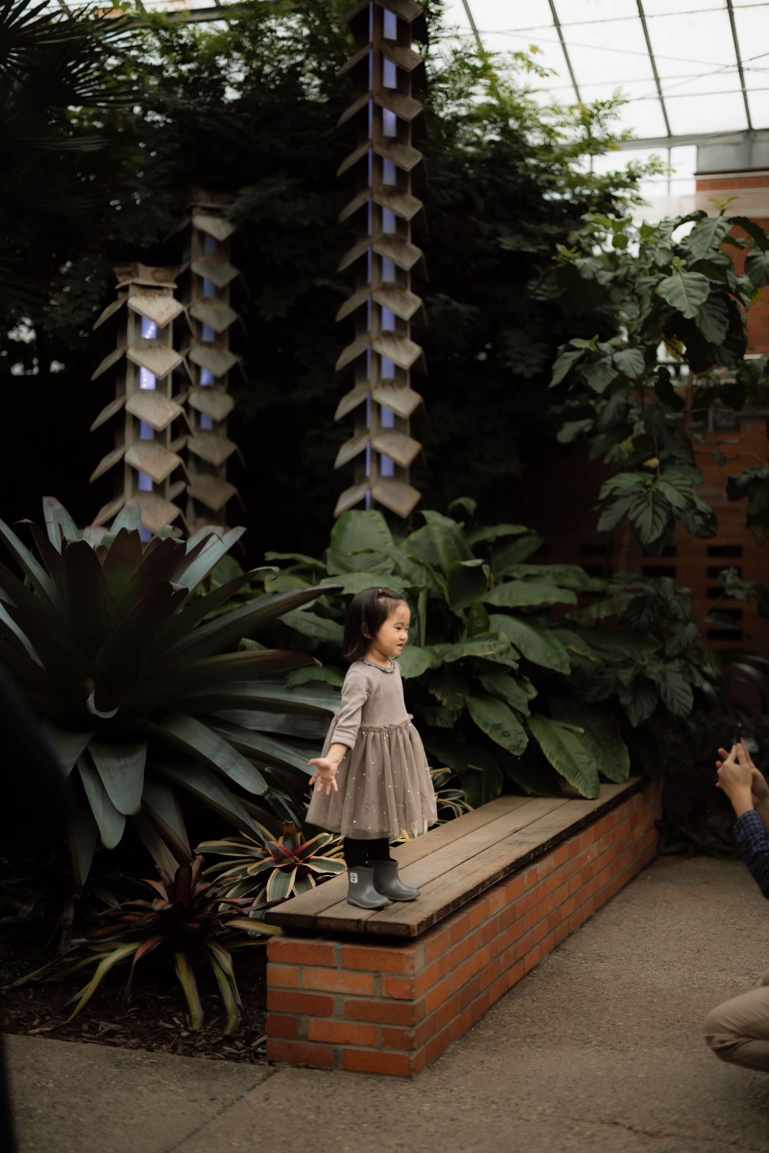 Young Asian girl in gray dress stands on brick planter in lush tropical greenhouse conservatory.