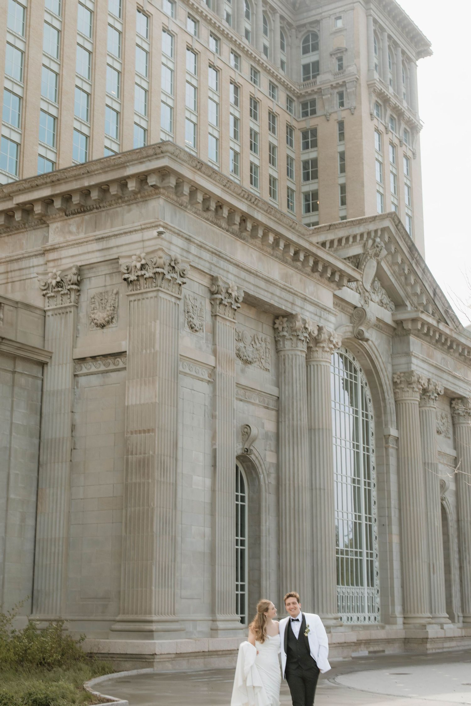 Bride and groom pose together outside a grand neoclassical building with tall columns on their wedding day.