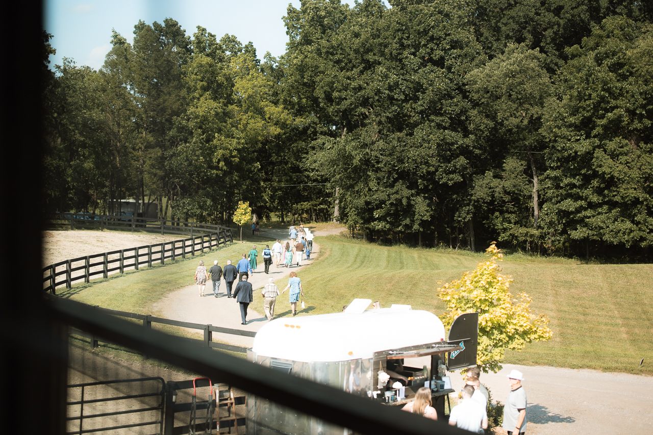 Aerial view of wedding guests gathering on outdoor path surrounded by lush green trees and white tent.