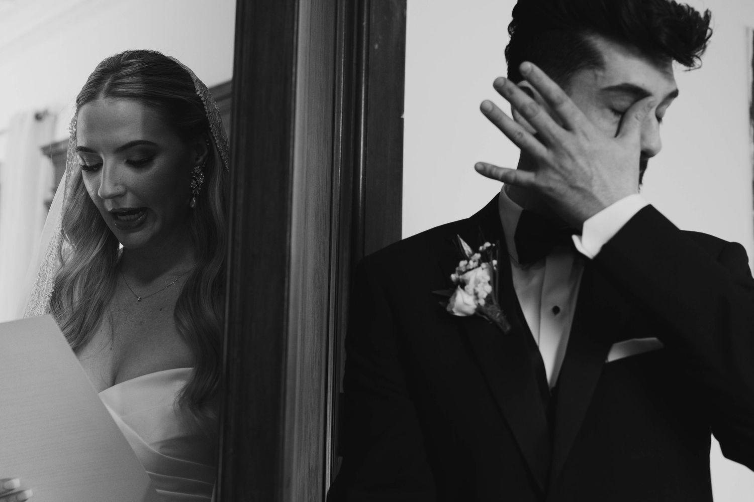 Black and white wedding photo of emotional groom covering face beside bride in white dress near window.