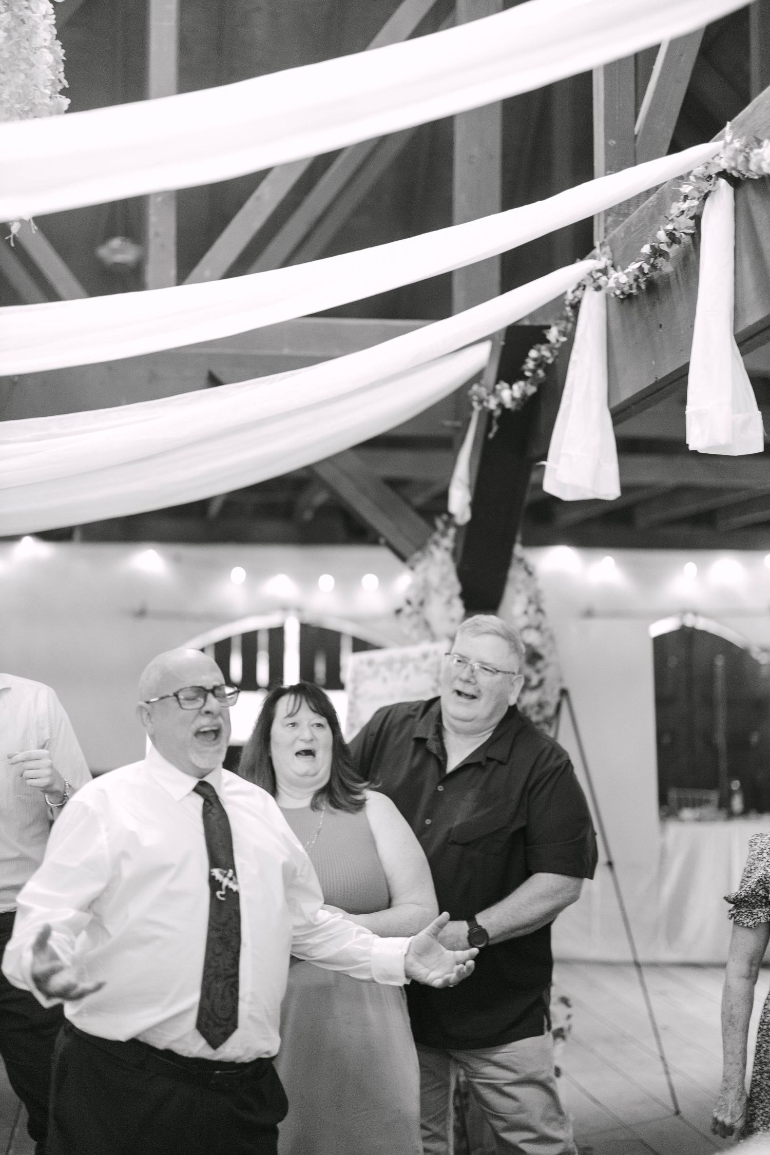 Black and white photo of three guests laughing and singing together at a wedding reception under draped fabric decorations.