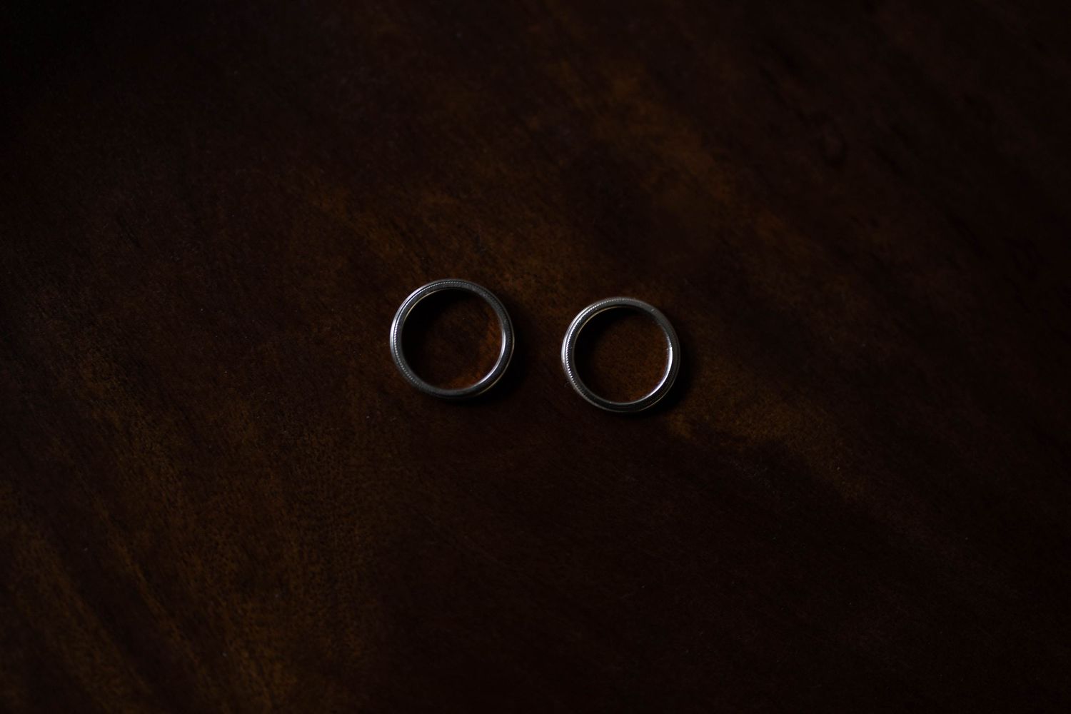 Two silver wedding rings sit on a dark wooden surface in dramatic lighting.