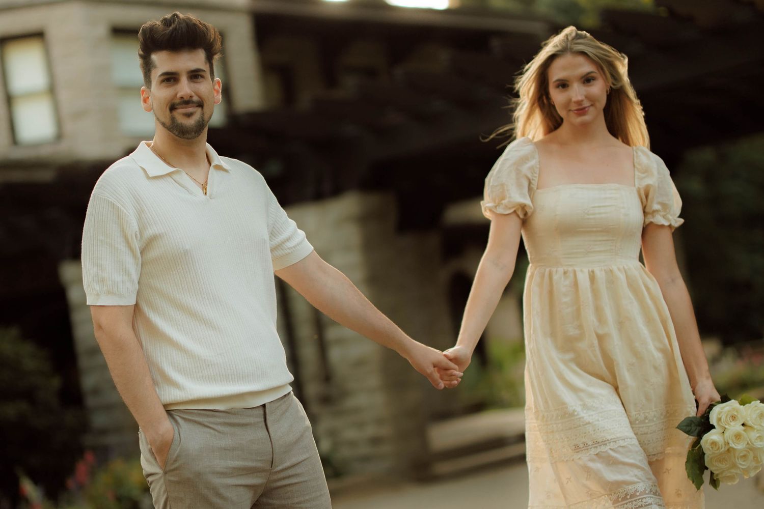 Couple holding hands outdoors, man in white polo and gray pants, woman in cream dress holding flowers.