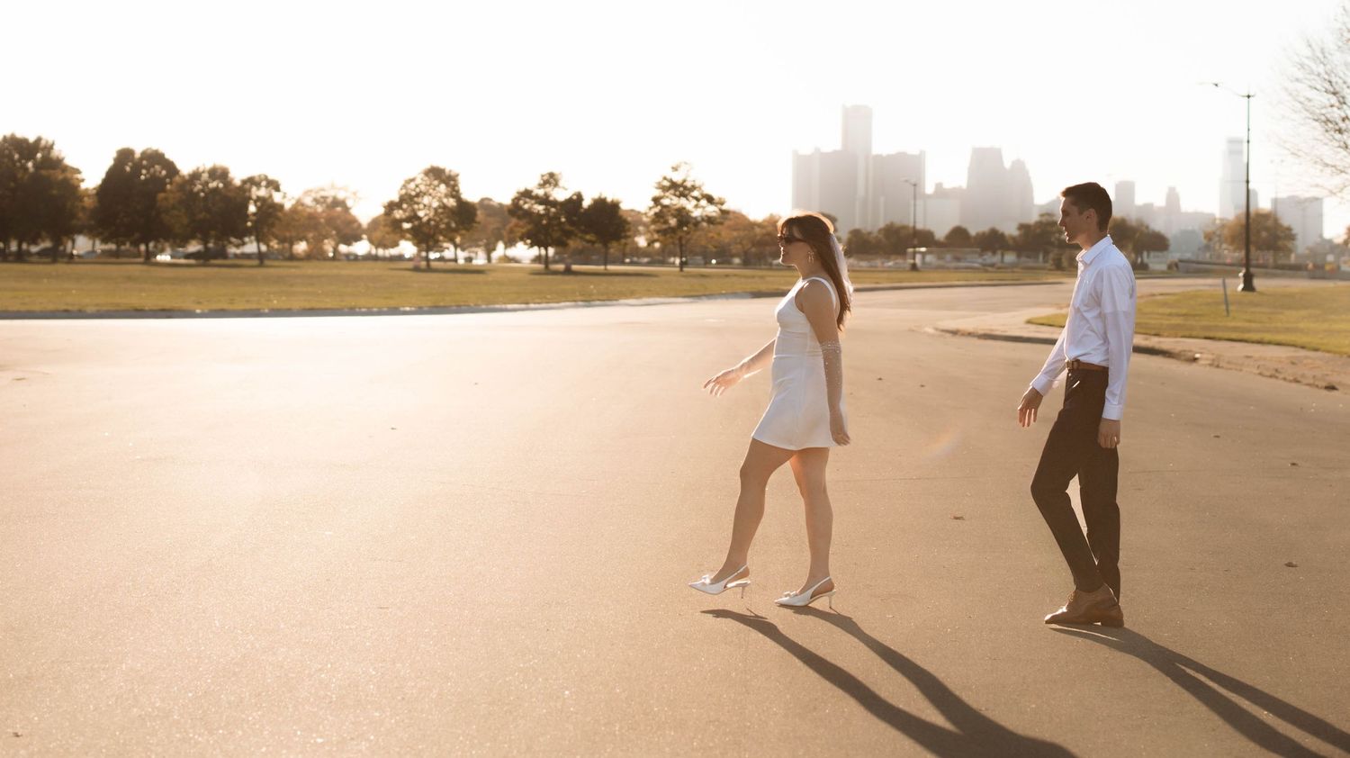 A couple walks hand in hand across an open paved area with a city skyline and trees in the warm golden sunlight.