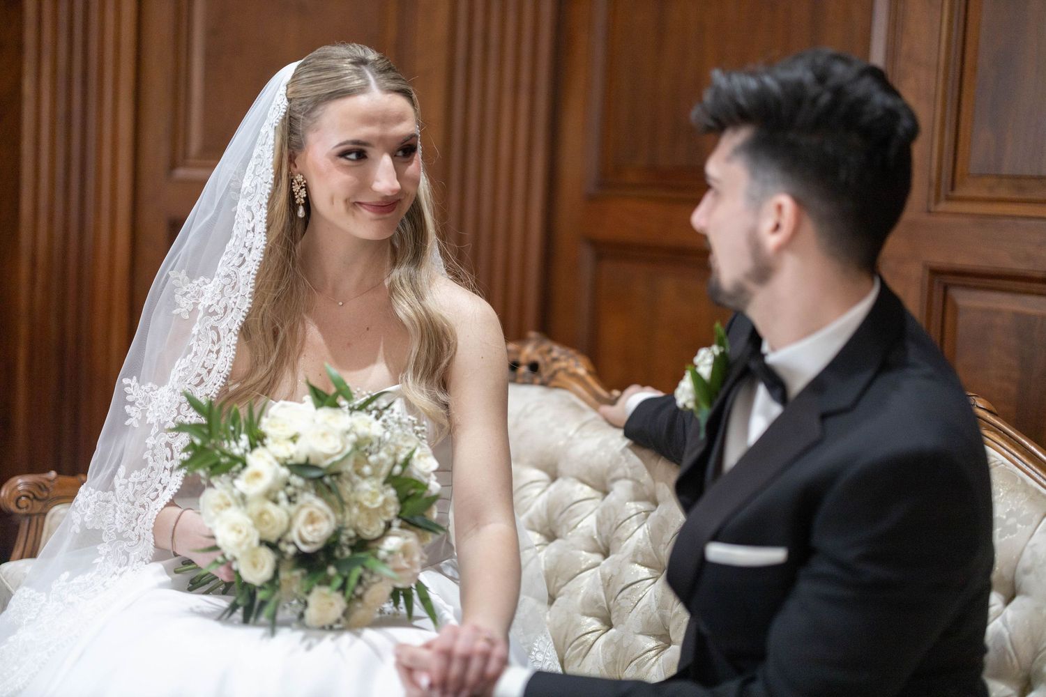 Bride in white gown and veil holds bouquet while smiling at groom in black tuxedo seated on elegant sofa.
