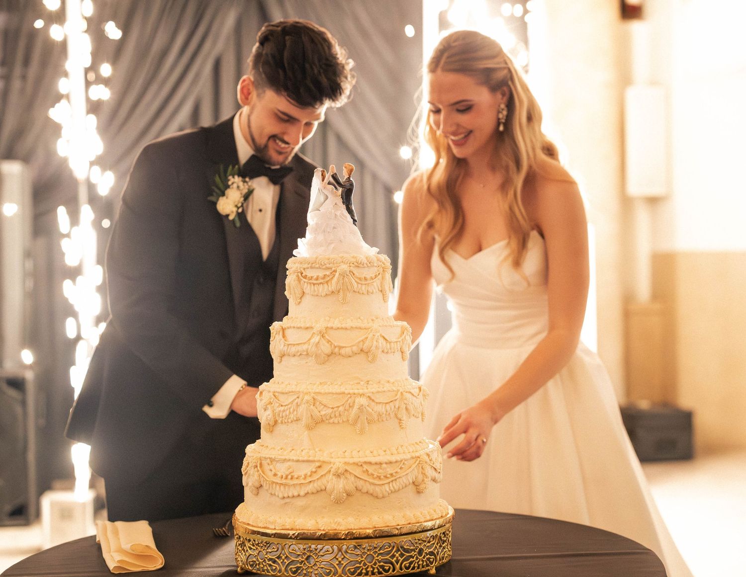 Bride and groom cutting elegant four-tier white wedding cake with sparkle backdrop at reception.