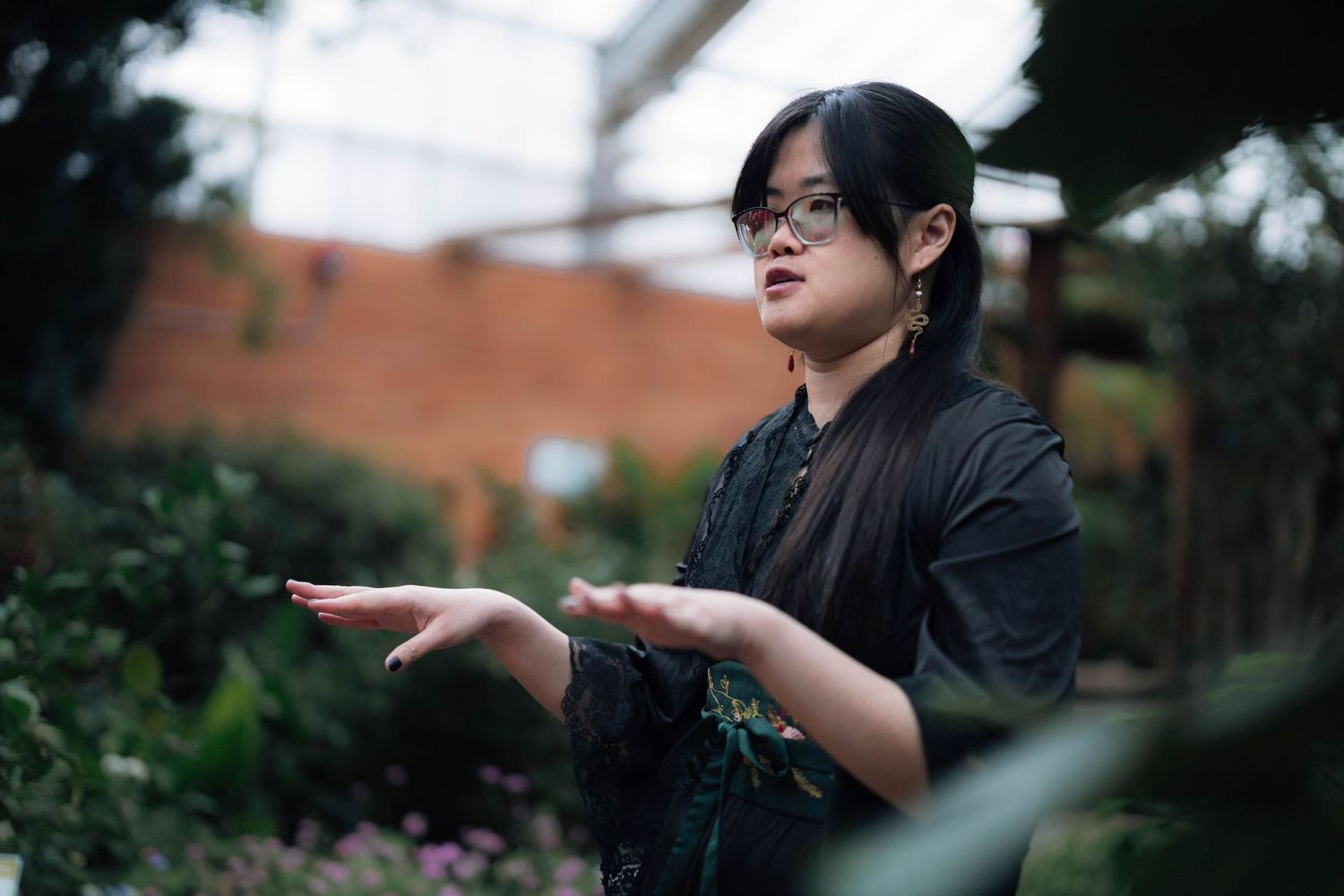 Asian woman with glasses gestures with open palms in lush garden setting.