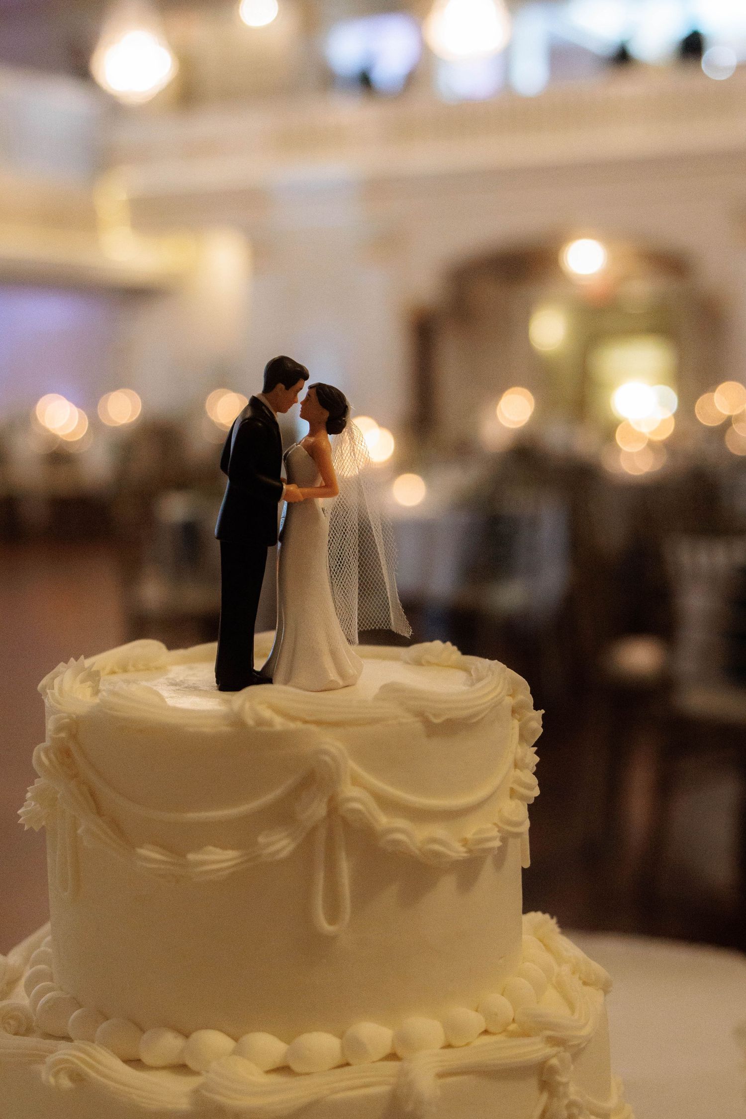 White wedding cake with bride and groom figurine topper in elegant reception hall with bokeh lights.