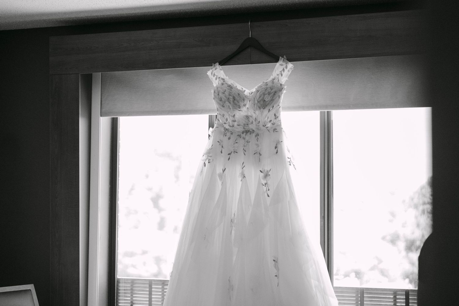 Elegant strapless wedding gown with beaded bodice hanging on a hanger in front of a window, black and white photo.