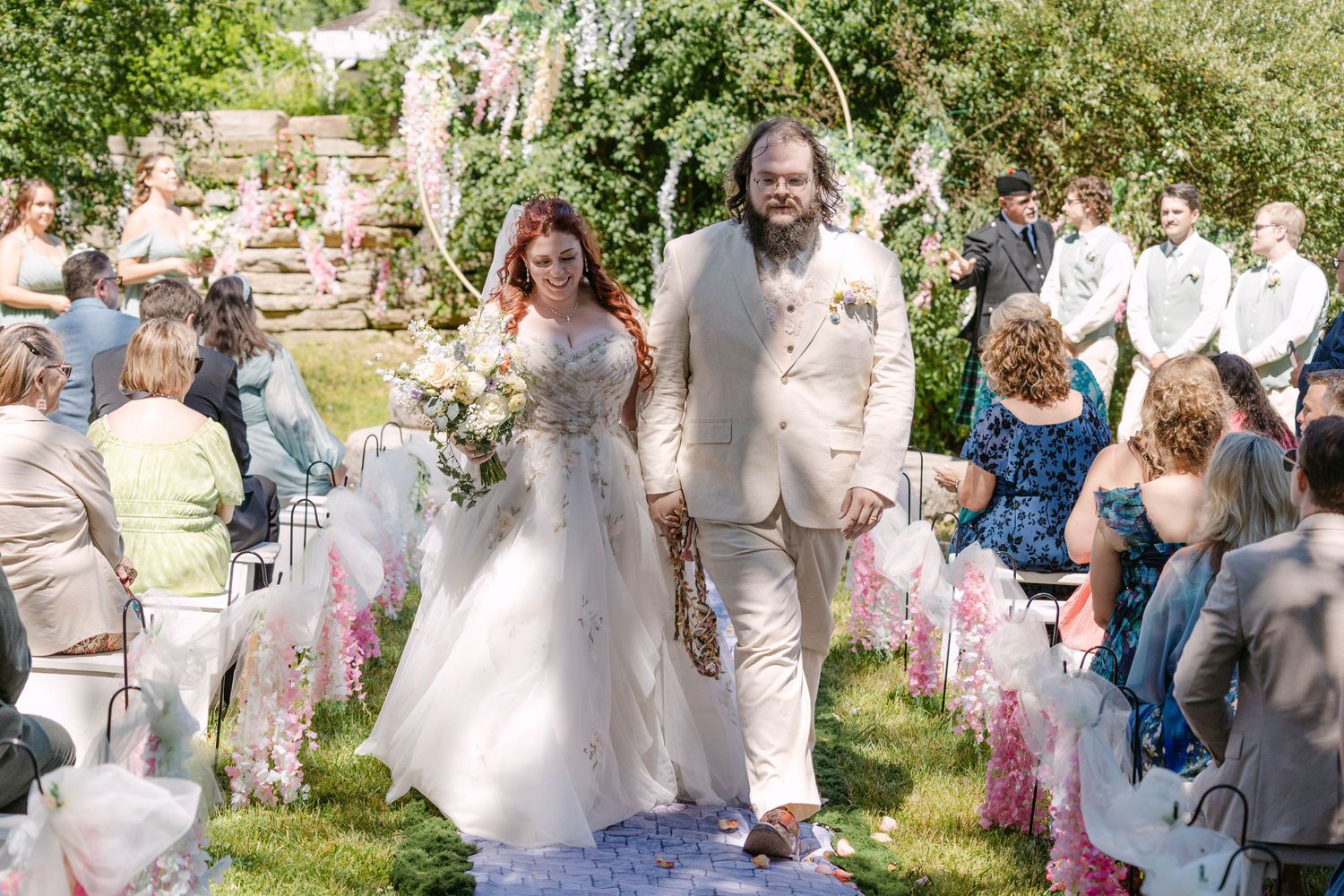 Bride and groom walk down outdoor wedding aisle, surrounded by seated guests and floral decorations on a sunny day.