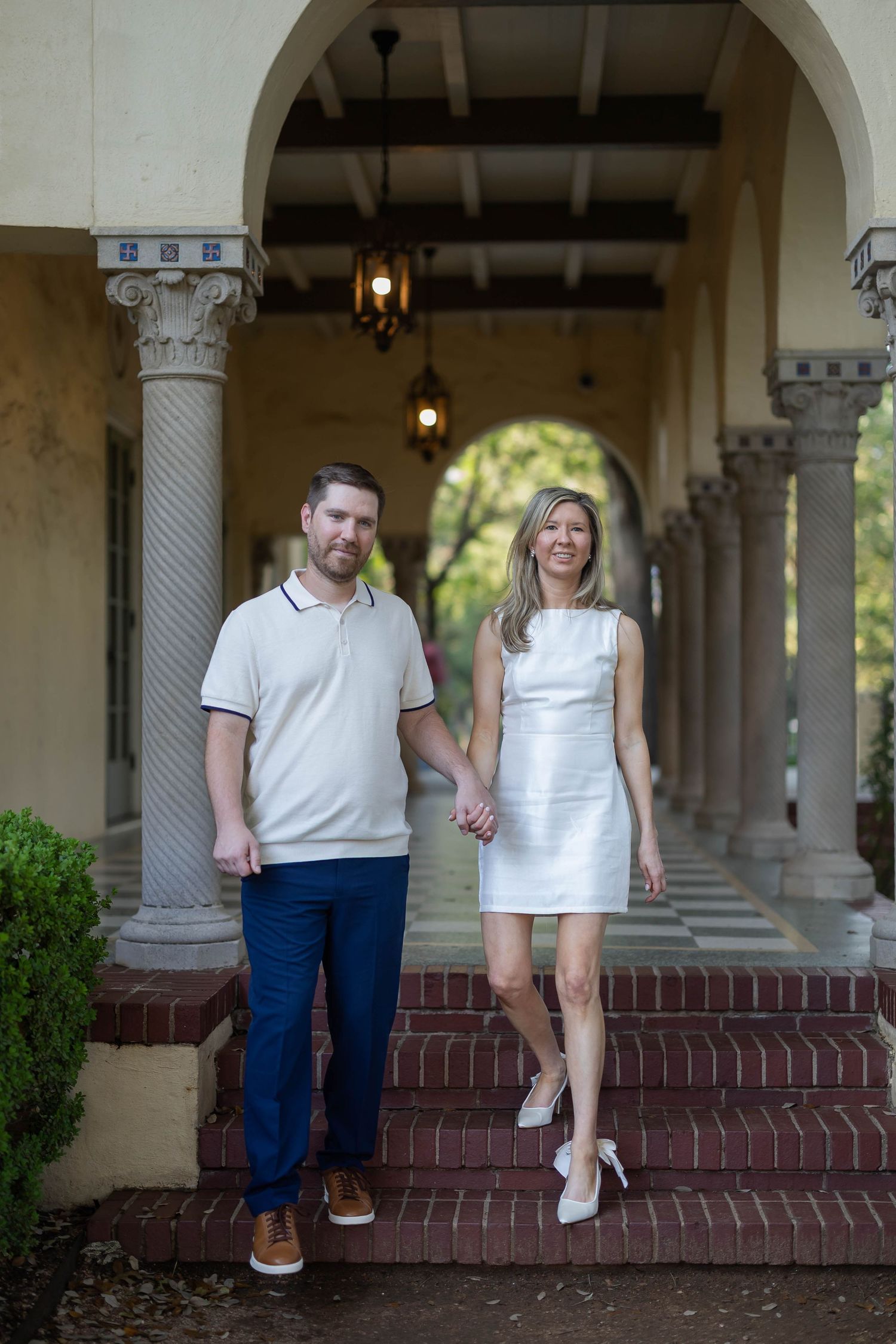 Couple holding hands on brick steps under arched colonnade with hanging lanterns.