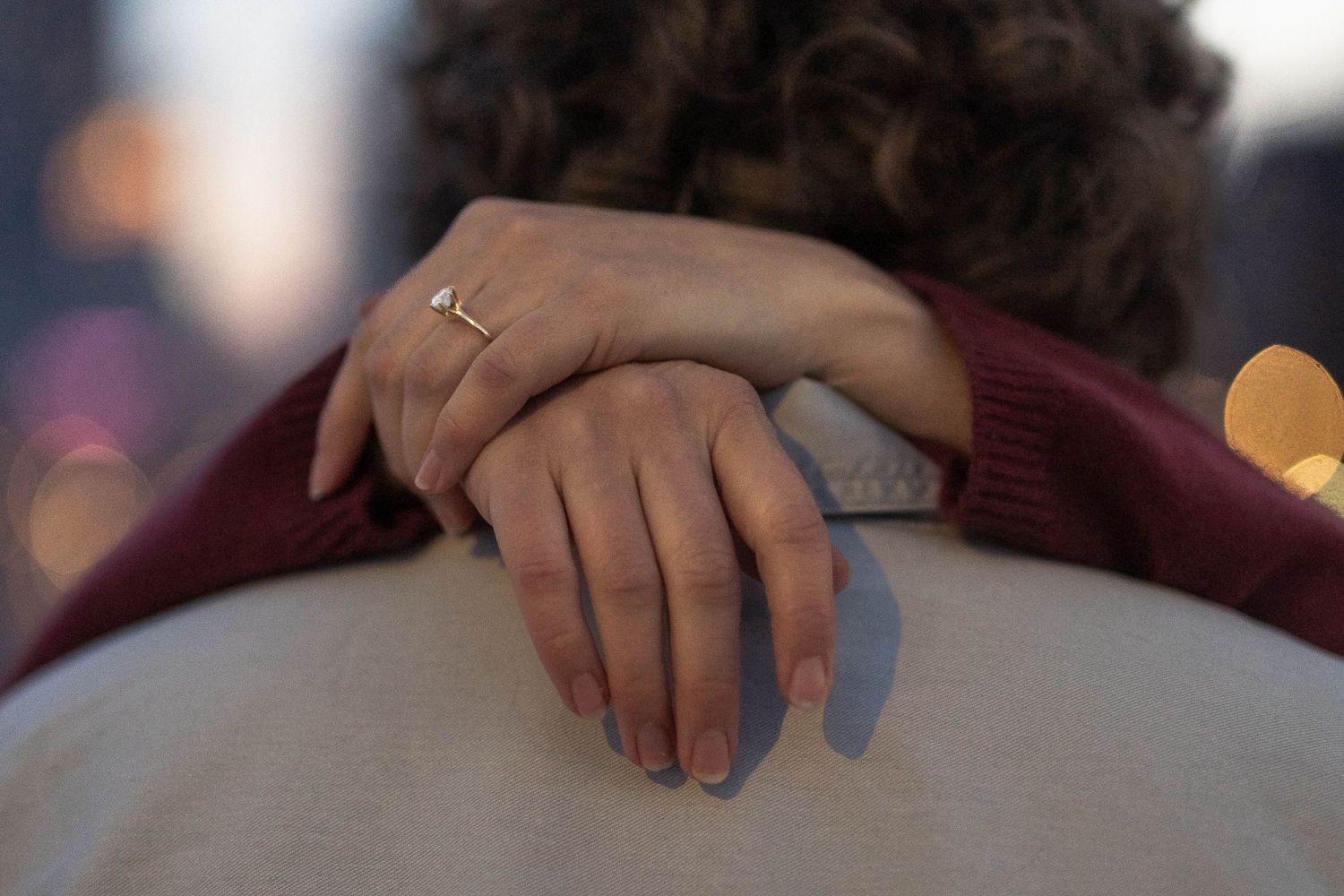 Close-up of a couple embracing, woman's hand with gold ring resting on man's shoulder in soft bokeh light.