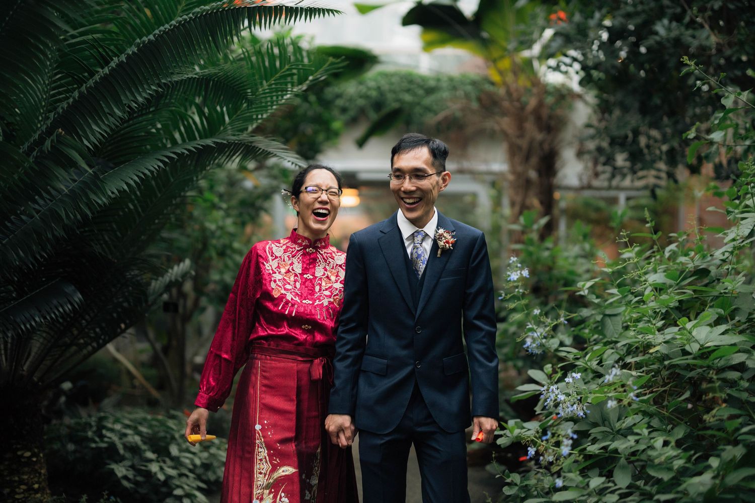 A couple in traditional formal attire poses together in a lush garden setting with ferns and greenery.