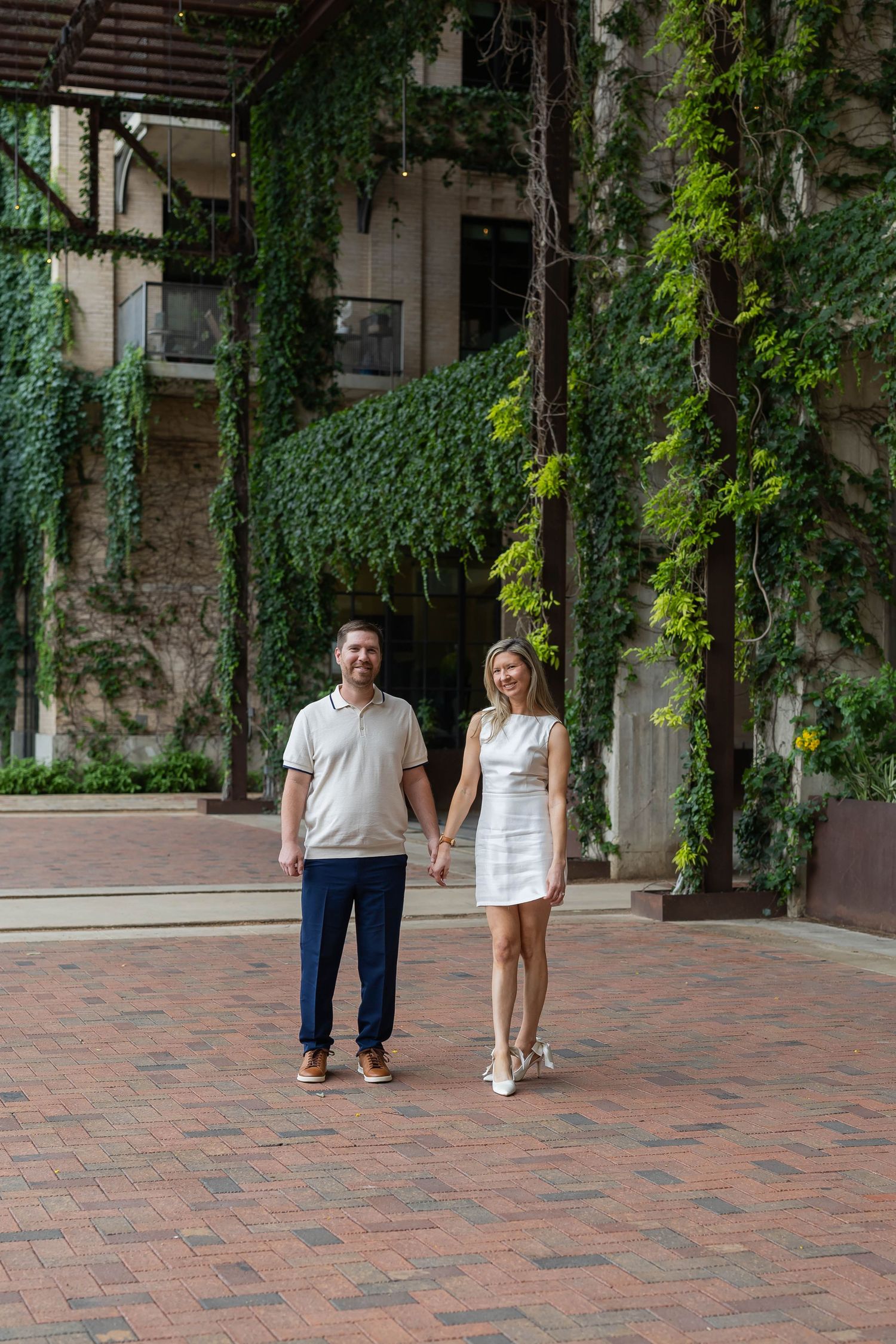 Couple holding hands and smiling in a brick courtyard surrounded by lush ivy-covered walls.
