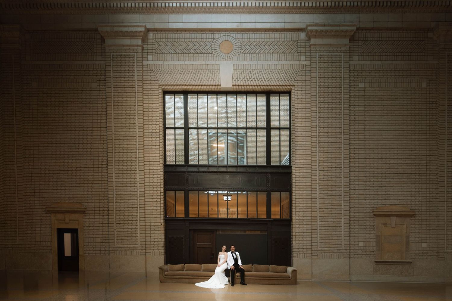 Wedding couple sitting together in a grand hall with large arched windows and ornate architecture.