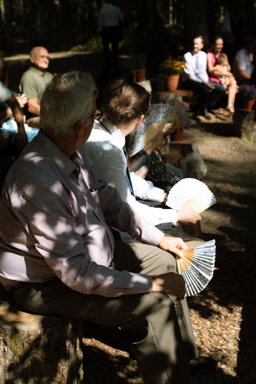 Wedding guests seated outdoors in dappled forest shade, using hand fans to stay cool during ceremony.