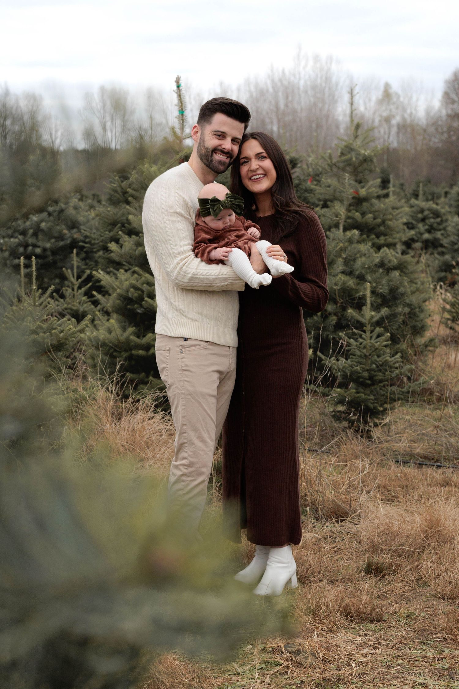 Happy couple holding baby at Christmas tree farm, woman in brown dress, man in cream sweater, surrounded by evergreen trees.