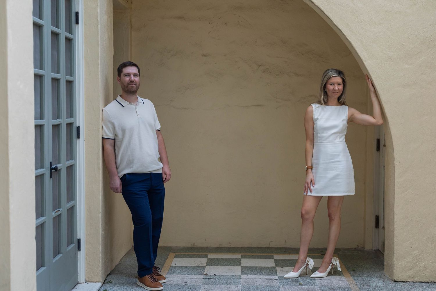 A man and woman stand apart in an elegant archway, he in blue jeans, she in a white mini dress and heels.