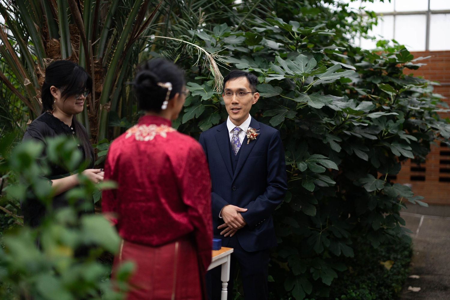 Couple in traditional Chinese wedding attire exchanging vows outdoors surrounded by lush tropical greenery.