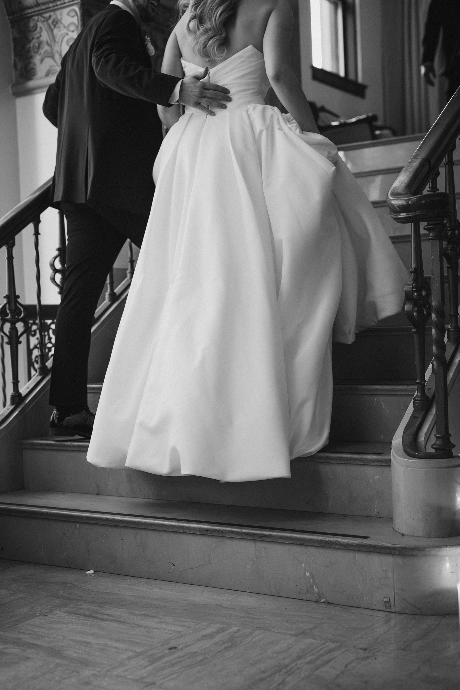 Black and white photo of bride and groom embracing on elegant staircase, bride in flowing white ball gown.