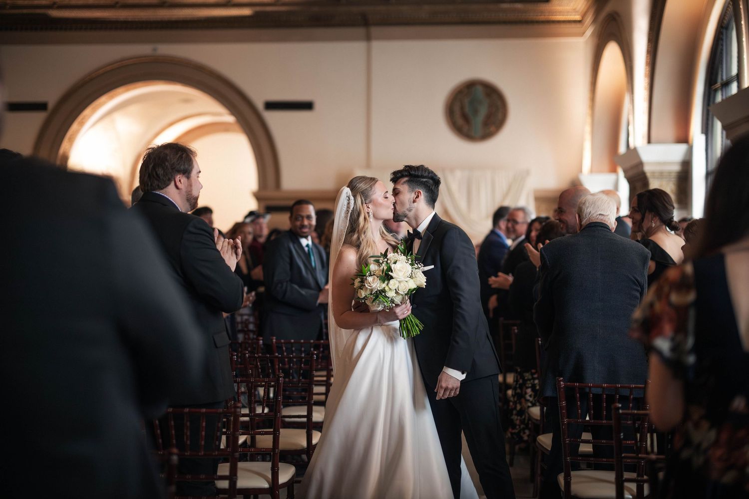 Bride and groom share first kiss at wedding ceremony aisle surrounded by guests in elegant venue.