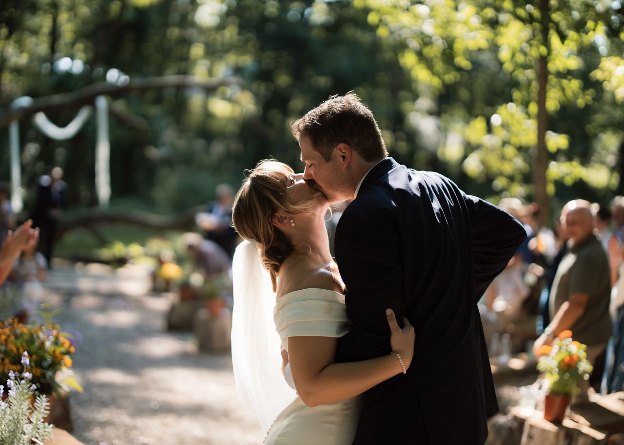 Bride and groom share first kiss at outdoor woodland wedding ceremony with guests watching.