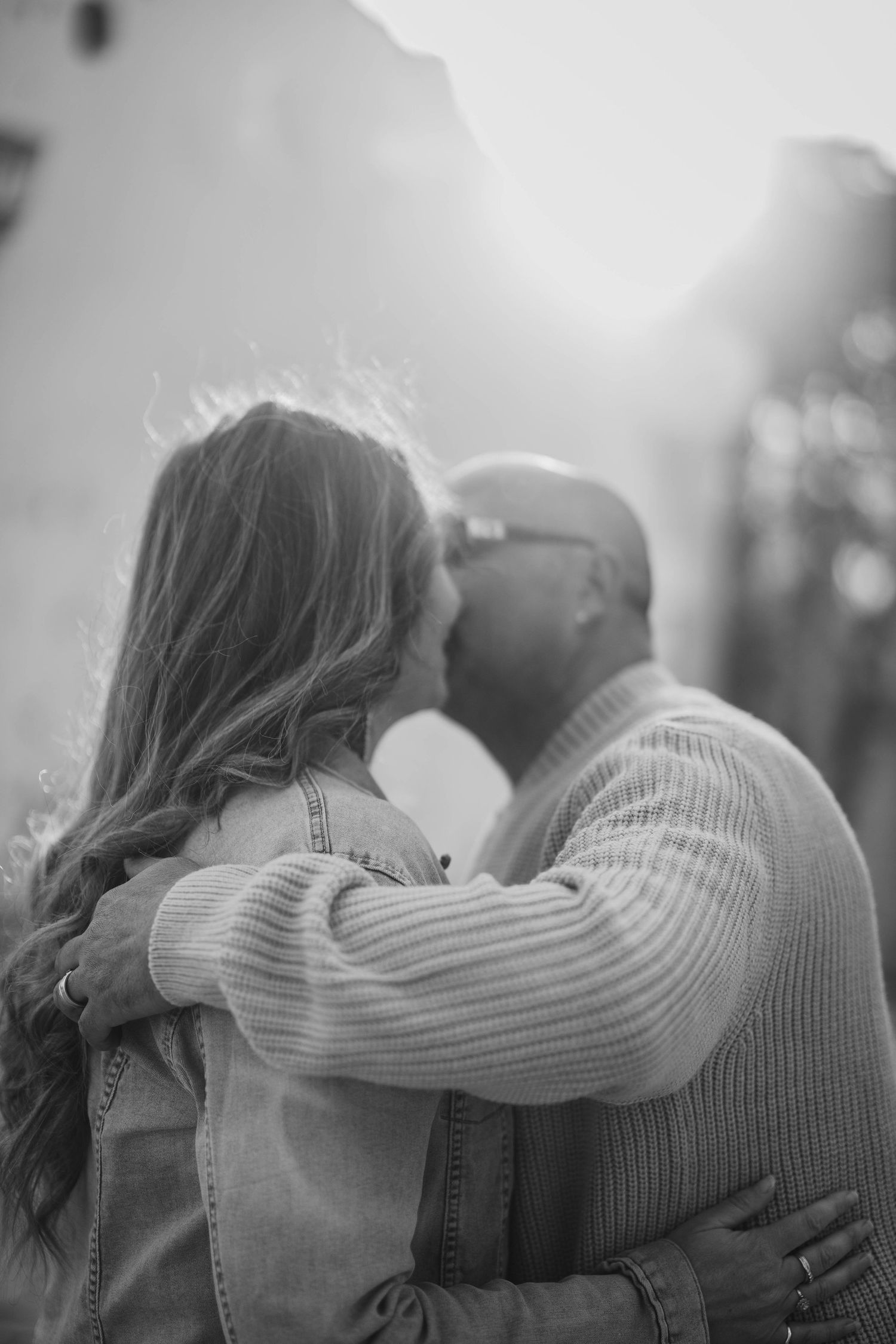 Black and white photo of a couple sharing a tender kiss, backlit by soft sunlight, woman with long hair embraced by bald man.