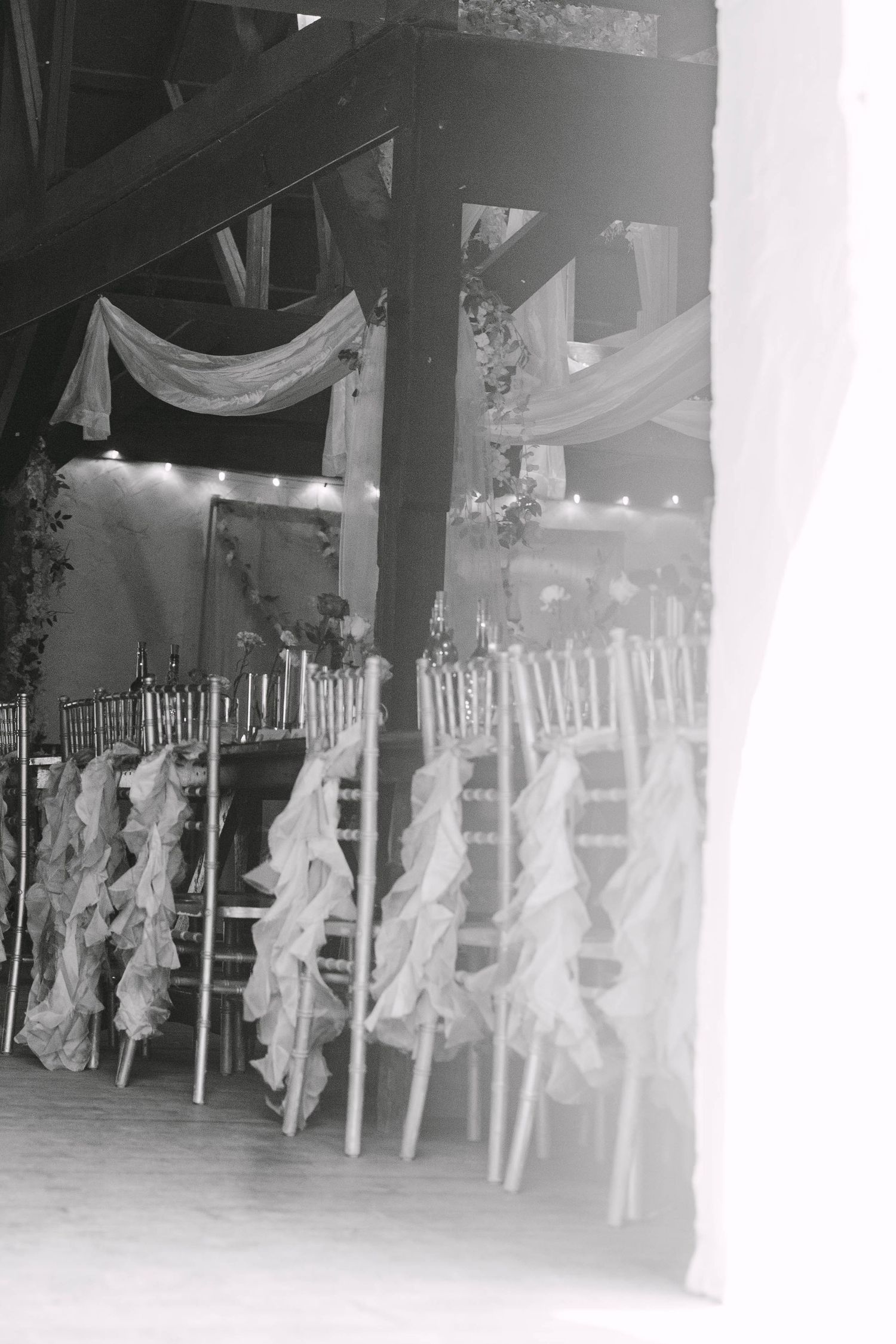 Black and white photo of elegant wedding ceremony chairs with ruffled sashes in a decorated barn venue with draped fabric.