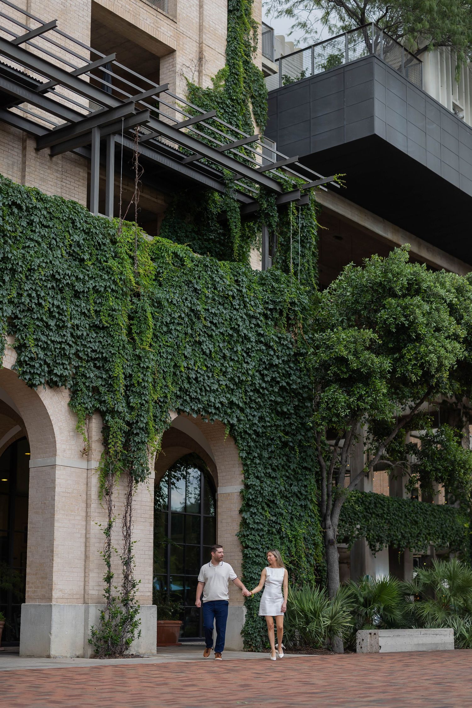 Couple holding hands walking past ivy-covered building with arched doorways.