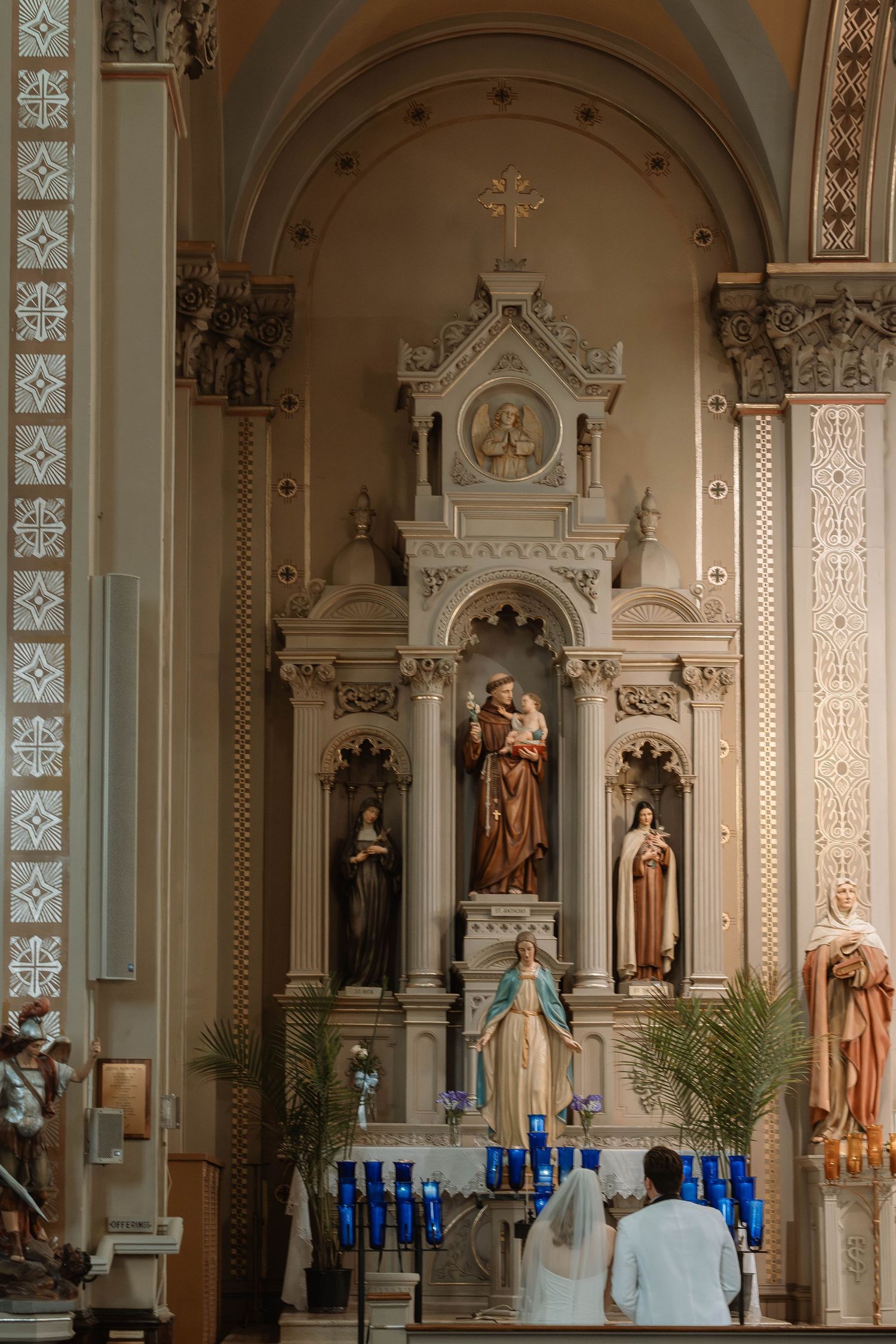 Ornate Catholic church altar with religious statues, blue candles, and a couple standing before the elaborate white reredos.