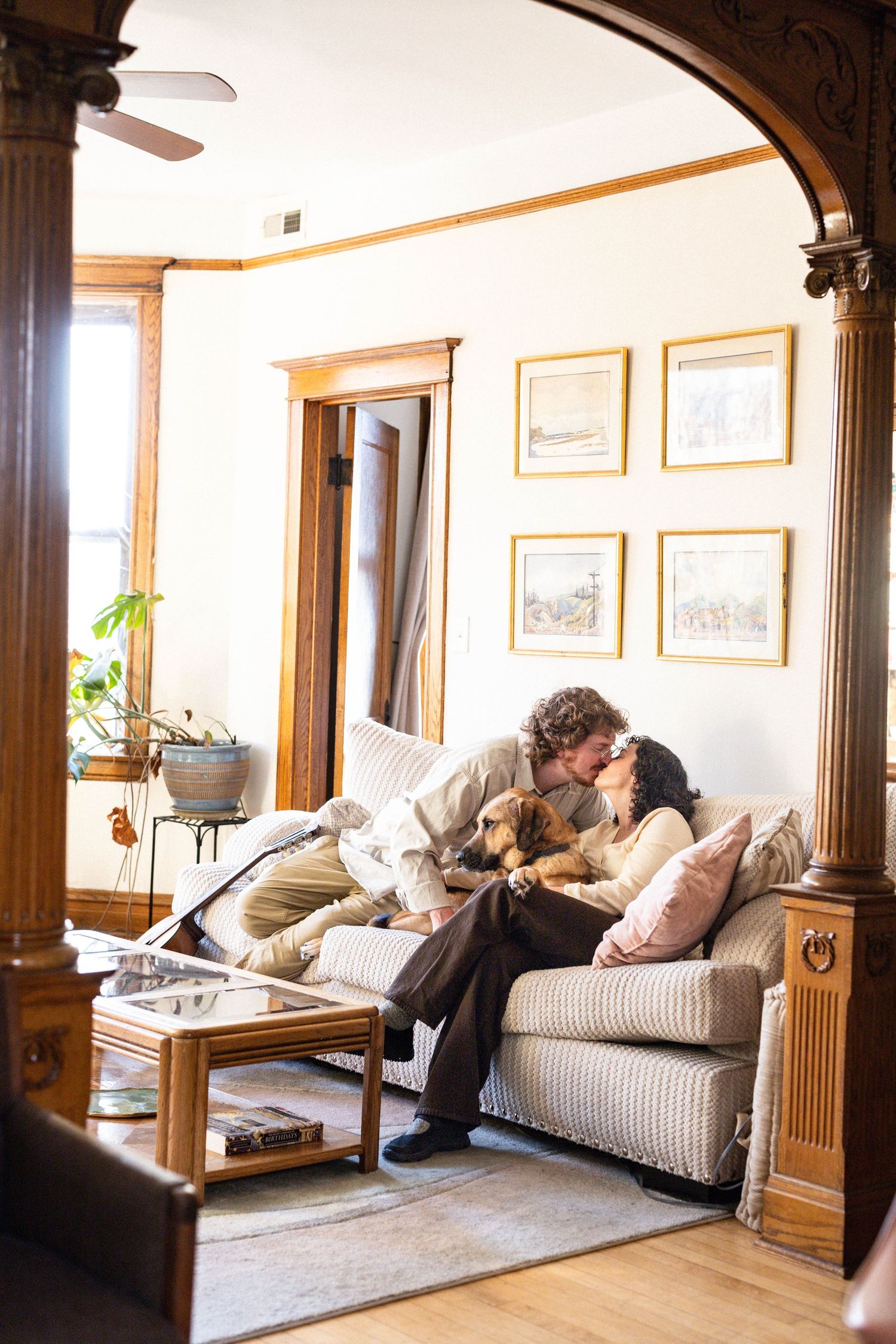 Couple kissing on couch with dog in cozy living room with arched doorway and framed art.