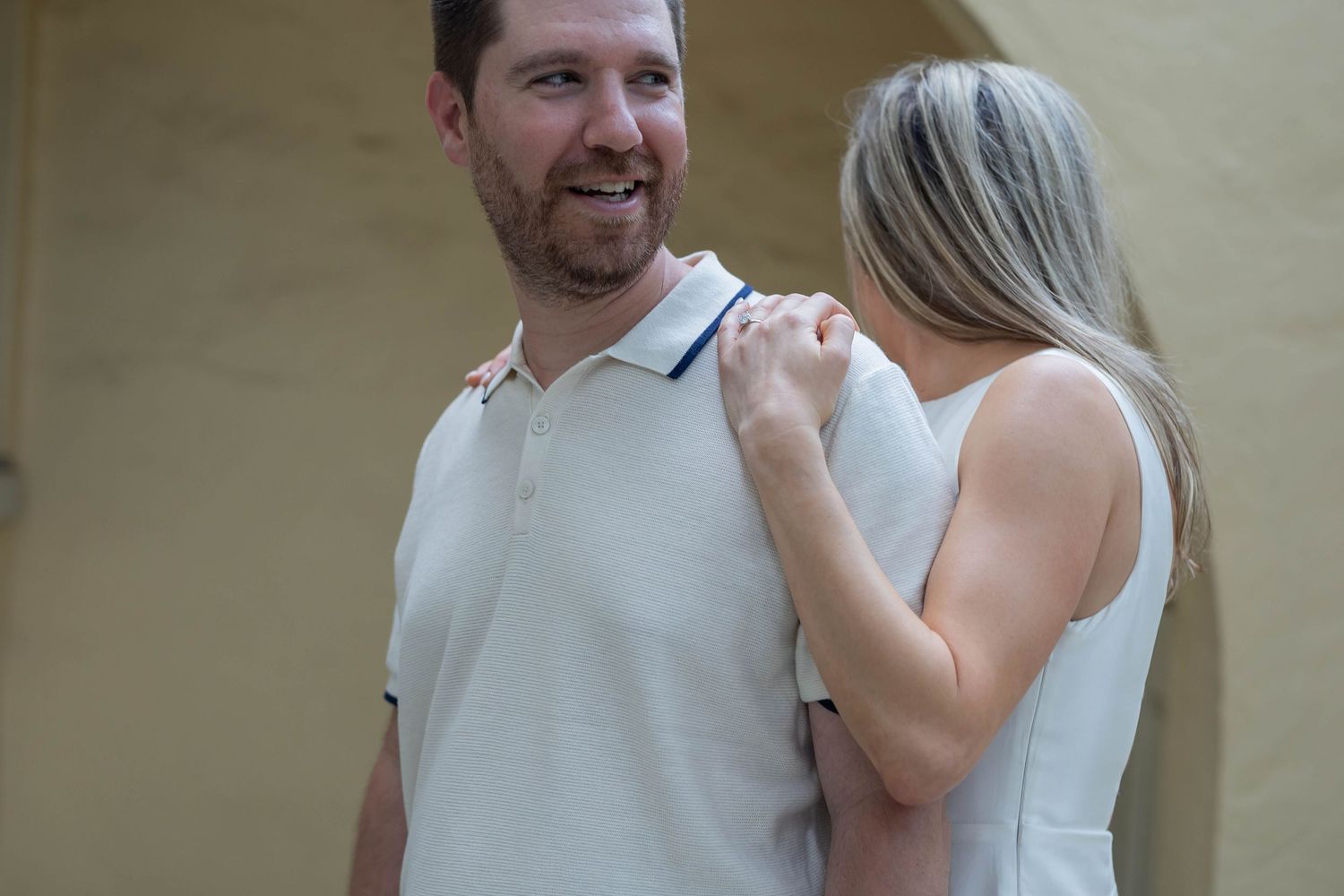 A smiling man in a white polo shirt gets his collar adjusted by a blonde woman in a sleeveless dress.