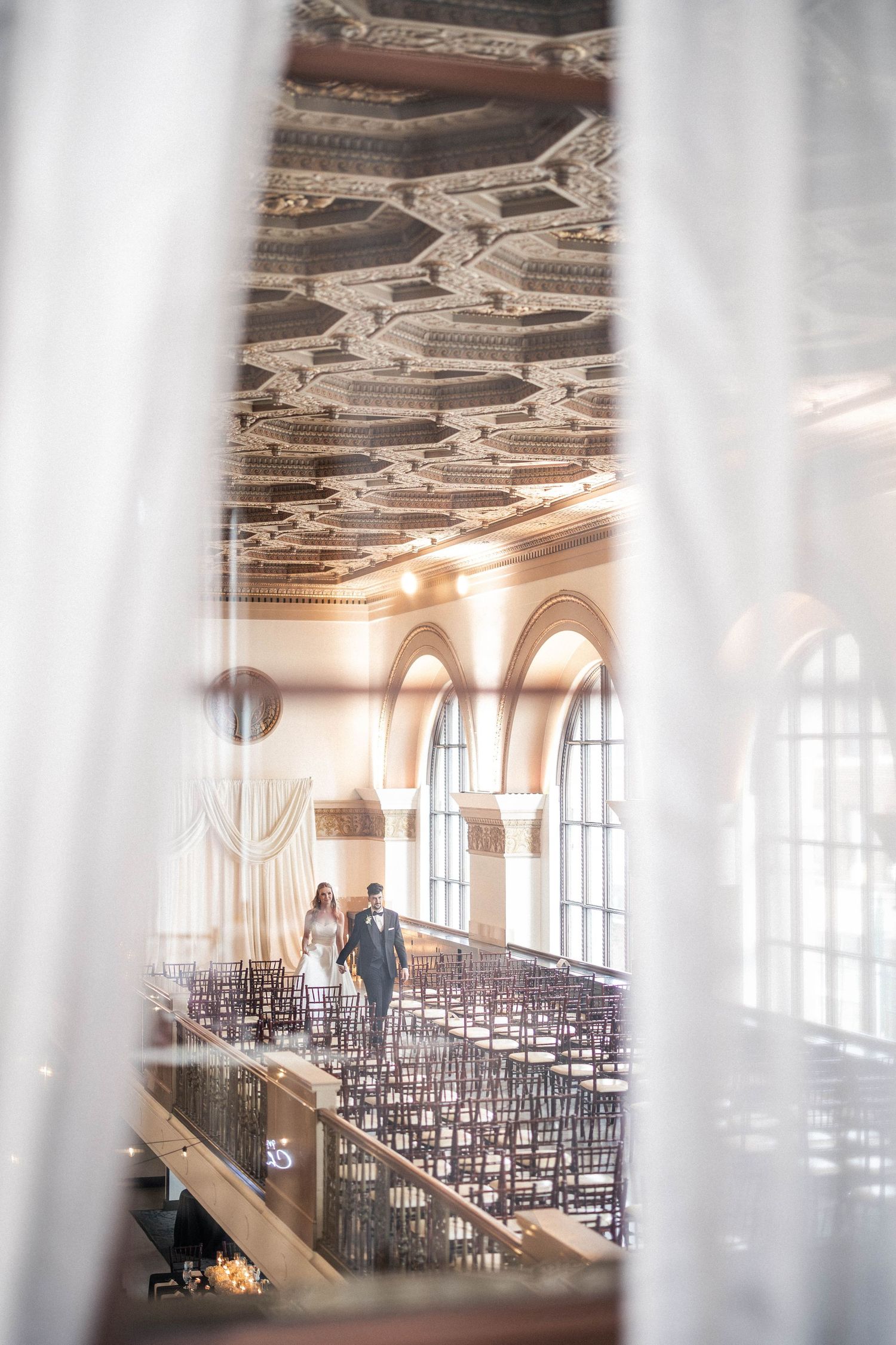 Bride and groom walk through an ornate wedding venue with arched windows and decorative ceiling.
