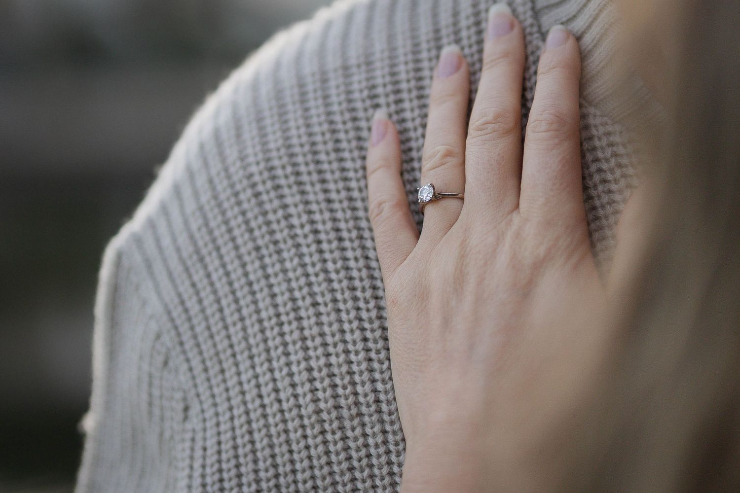 A close-up of a hand wearing a diamond solitaire engagement ring resting on a gray knit sweater.