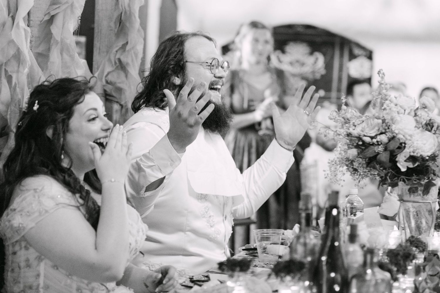 Black and white wedding reception photo of laughing bride and groom clapping at a decorated table.